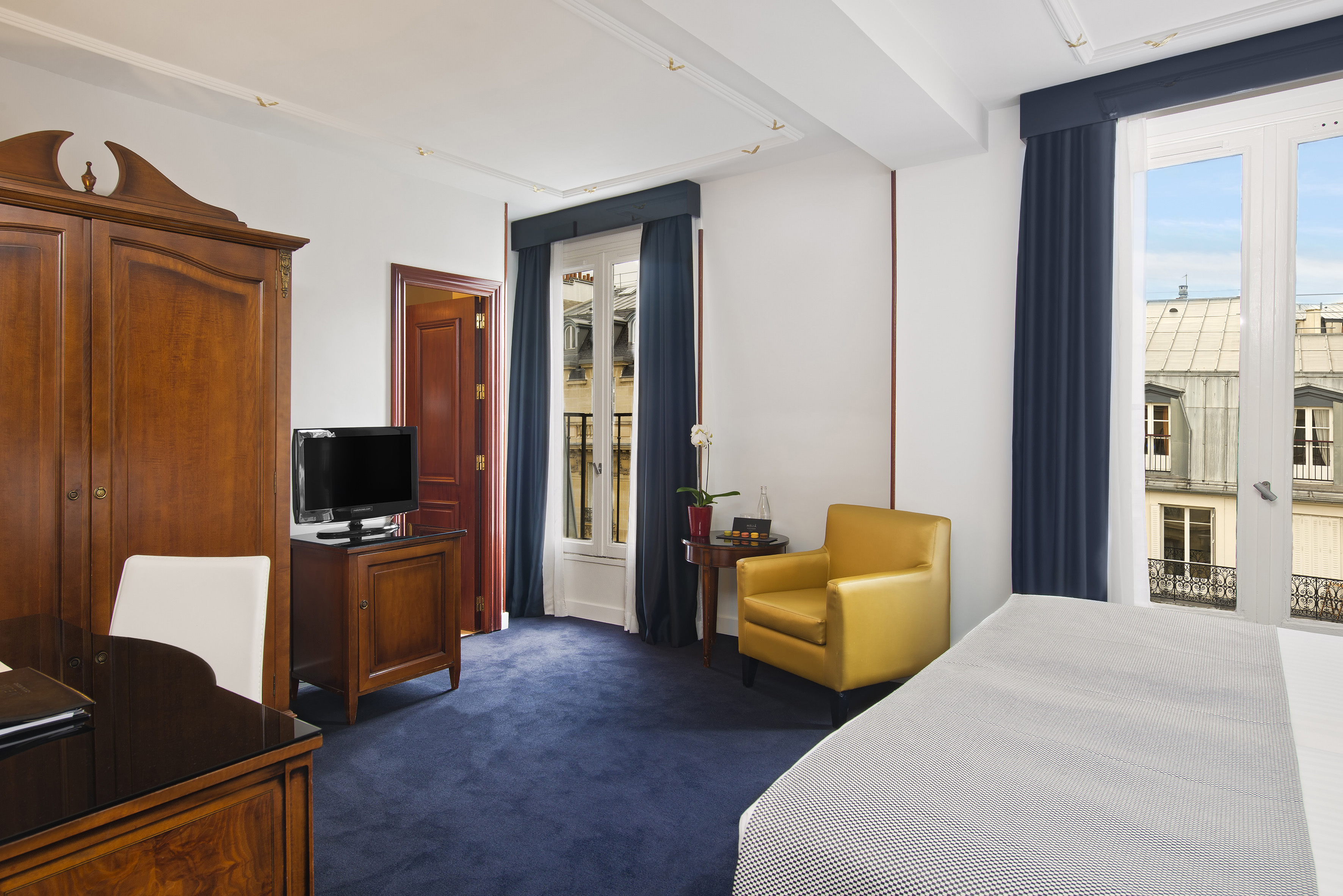Parisian hotel room: blue carpet, wooden wardrobe, yellow armchair, city view.