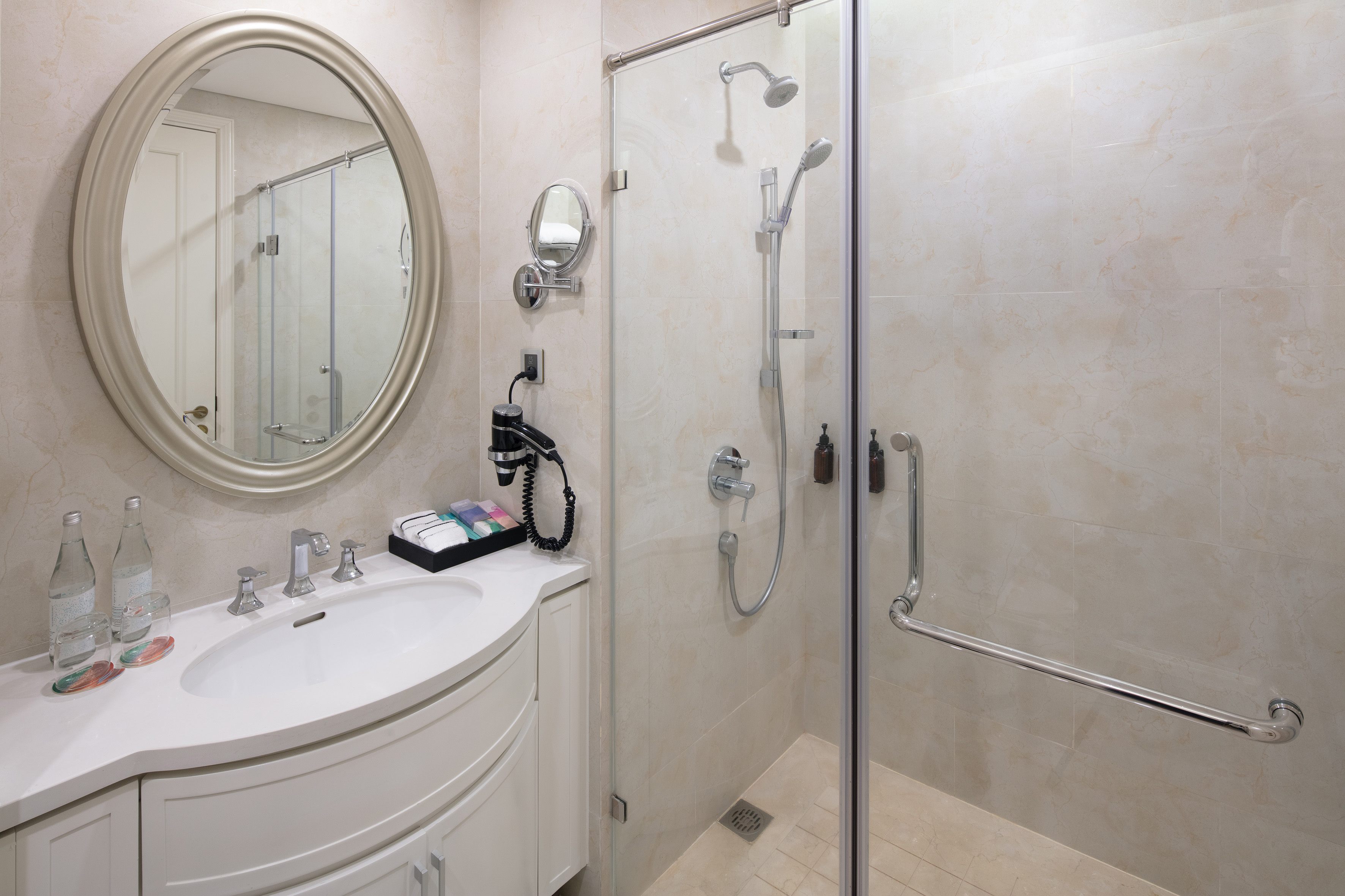 Modern bathroom with white vanity, oval mirror, glass shower, and beige tiles.