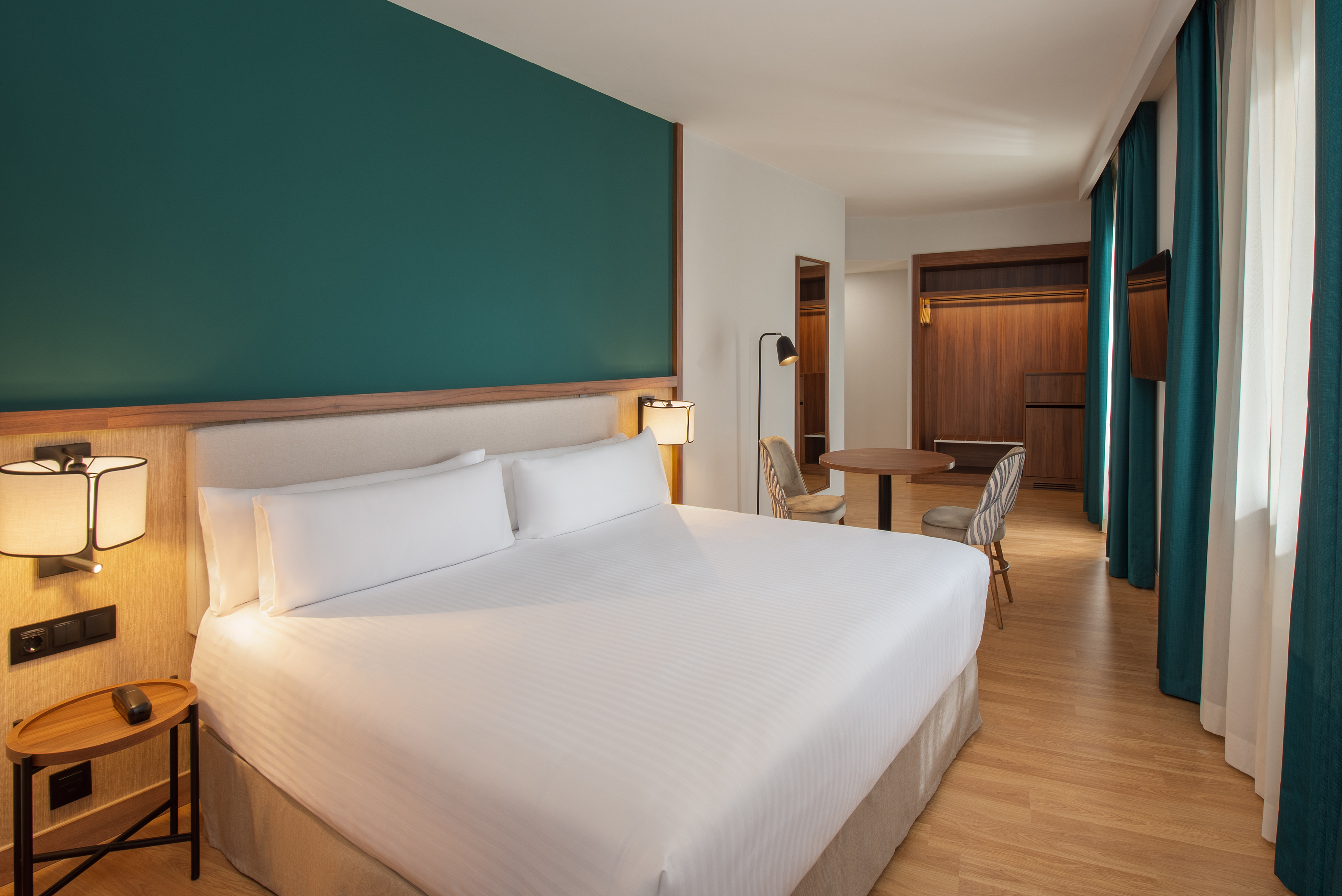 Modern hotel room with large bed, teal accent wall, and wooden furnishings.