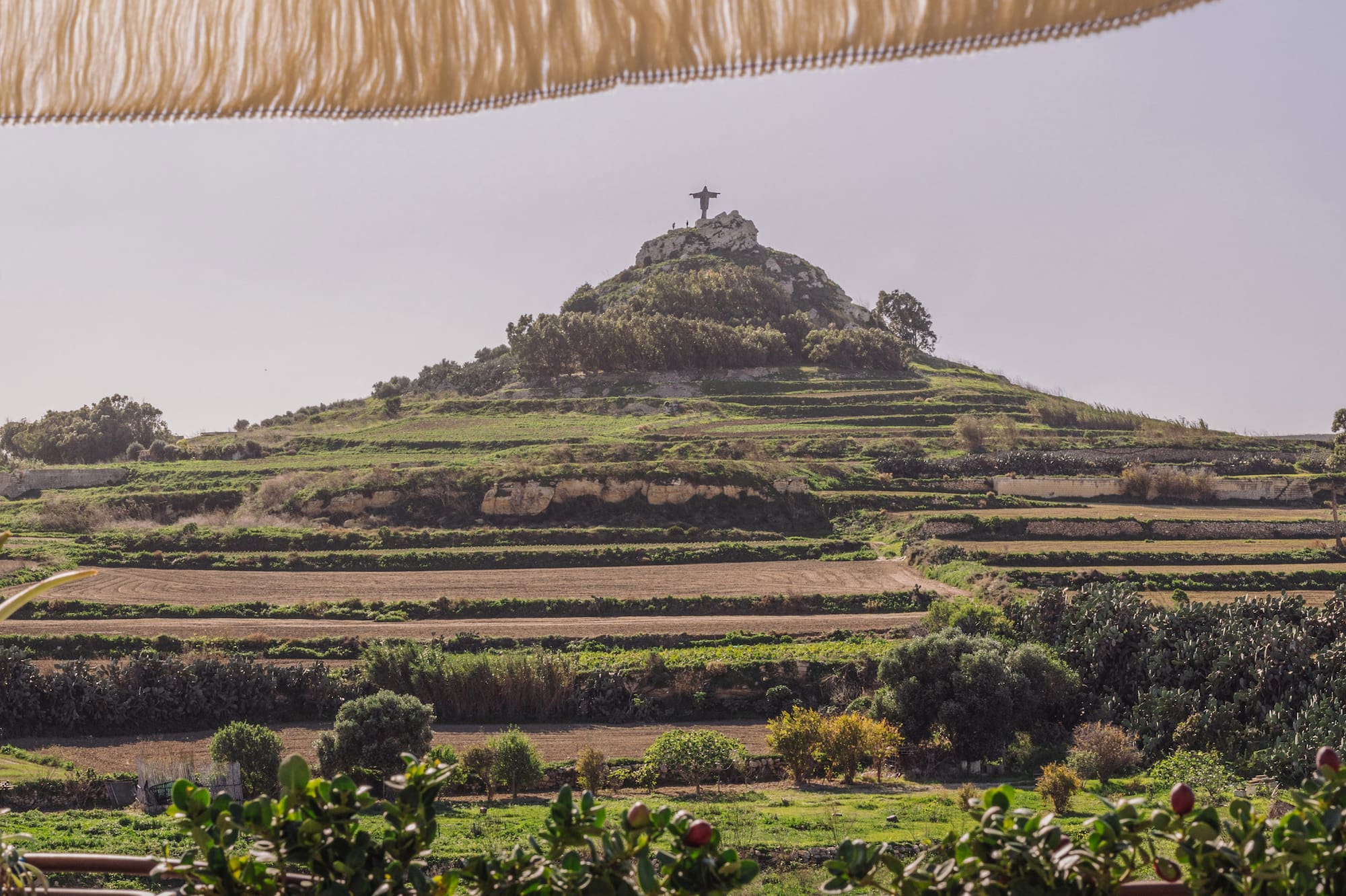 Fringed umbrella, planters, terraced fields leading to Christ statue on hill.