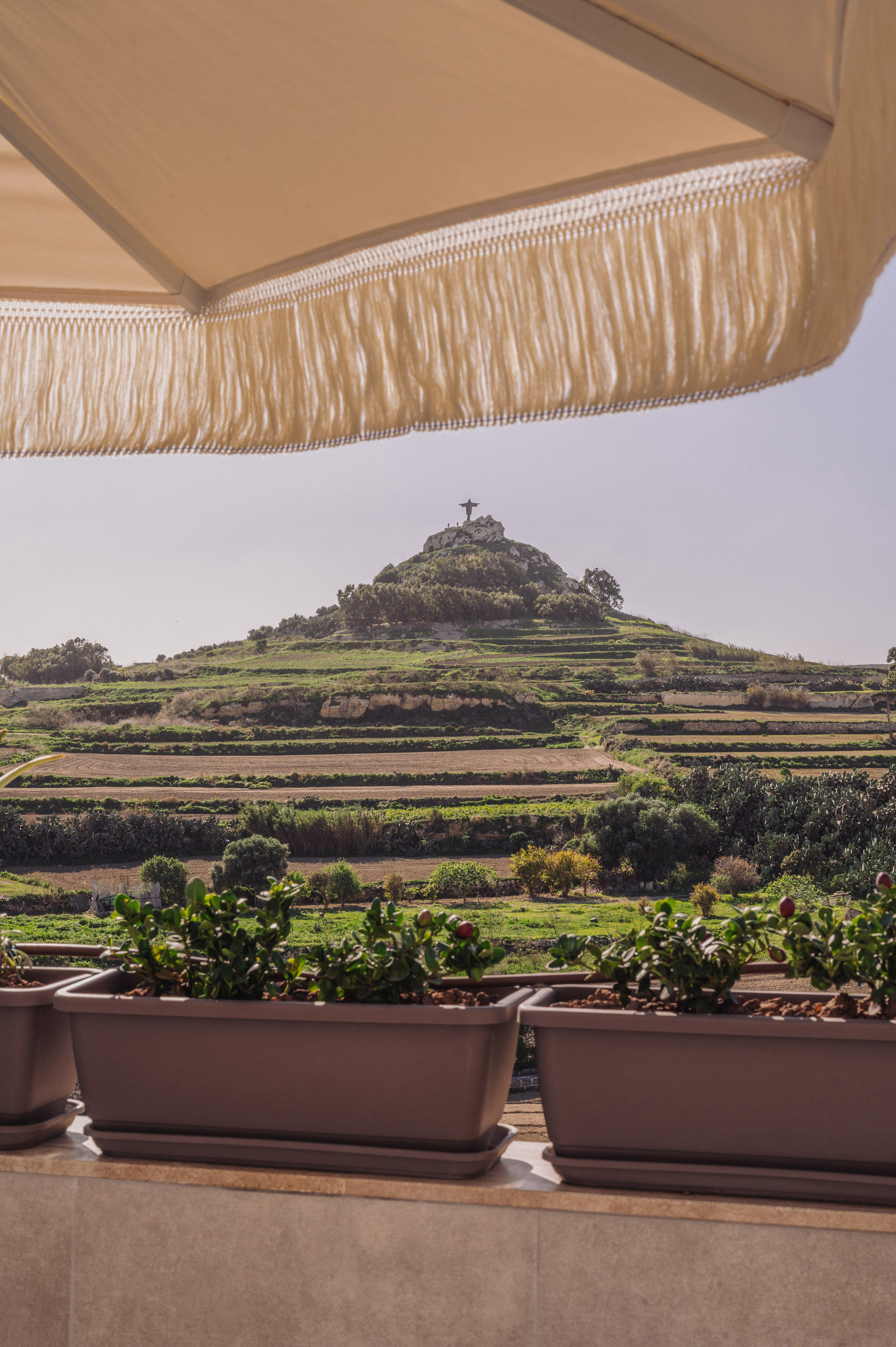 Fringed umbrella, planters, terraced fields leading to Christ statue on hill.