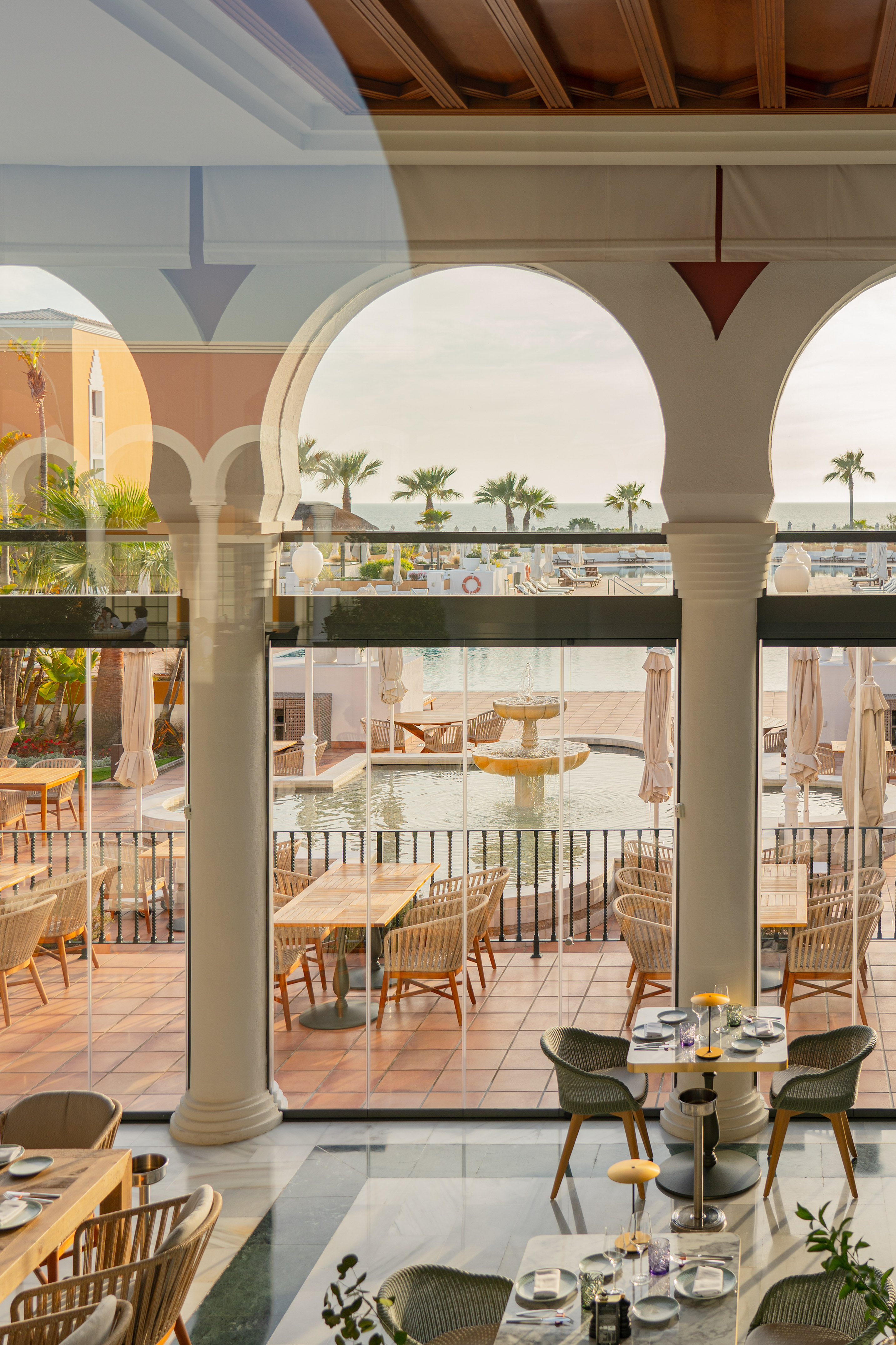 Hotel restaurant with arched windows framing a terrace, pool, ocean, and palms.