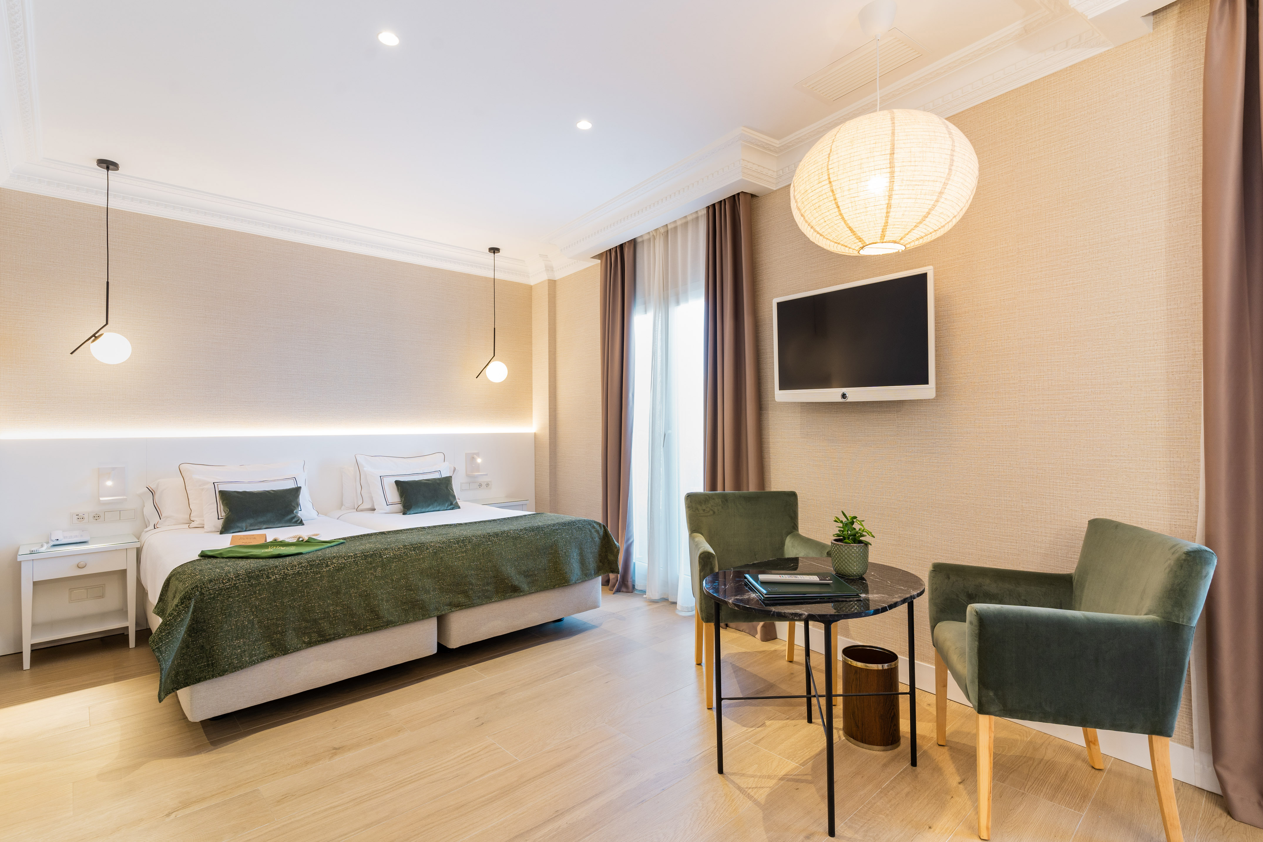 Modern hotel bedroom with twin beds, green decor, wall TV, and seating area.