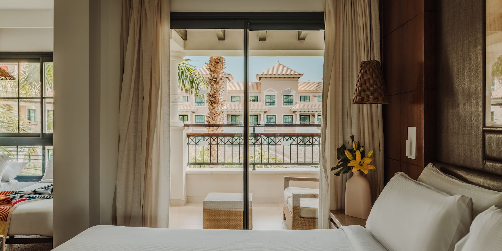 a bed with white sheets and a vase of flowers outside a glass door