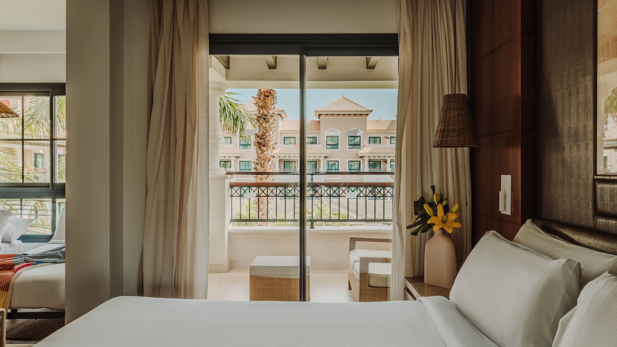 a bed with white sheets and a vase of flowers outside a glass door