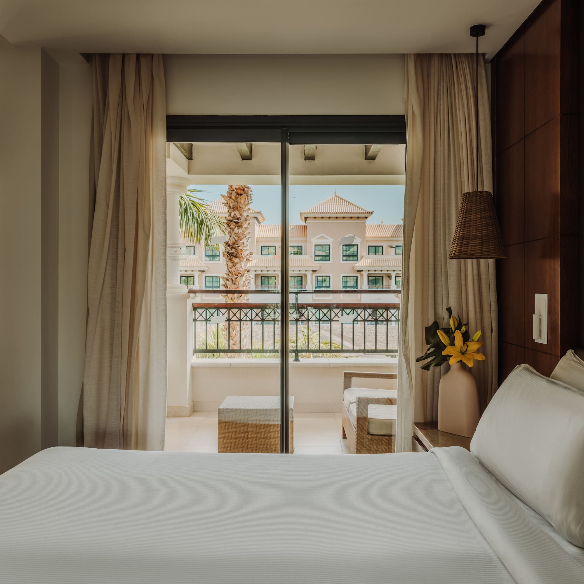 a bed with white sheets and a vase of flowers outside a glass door