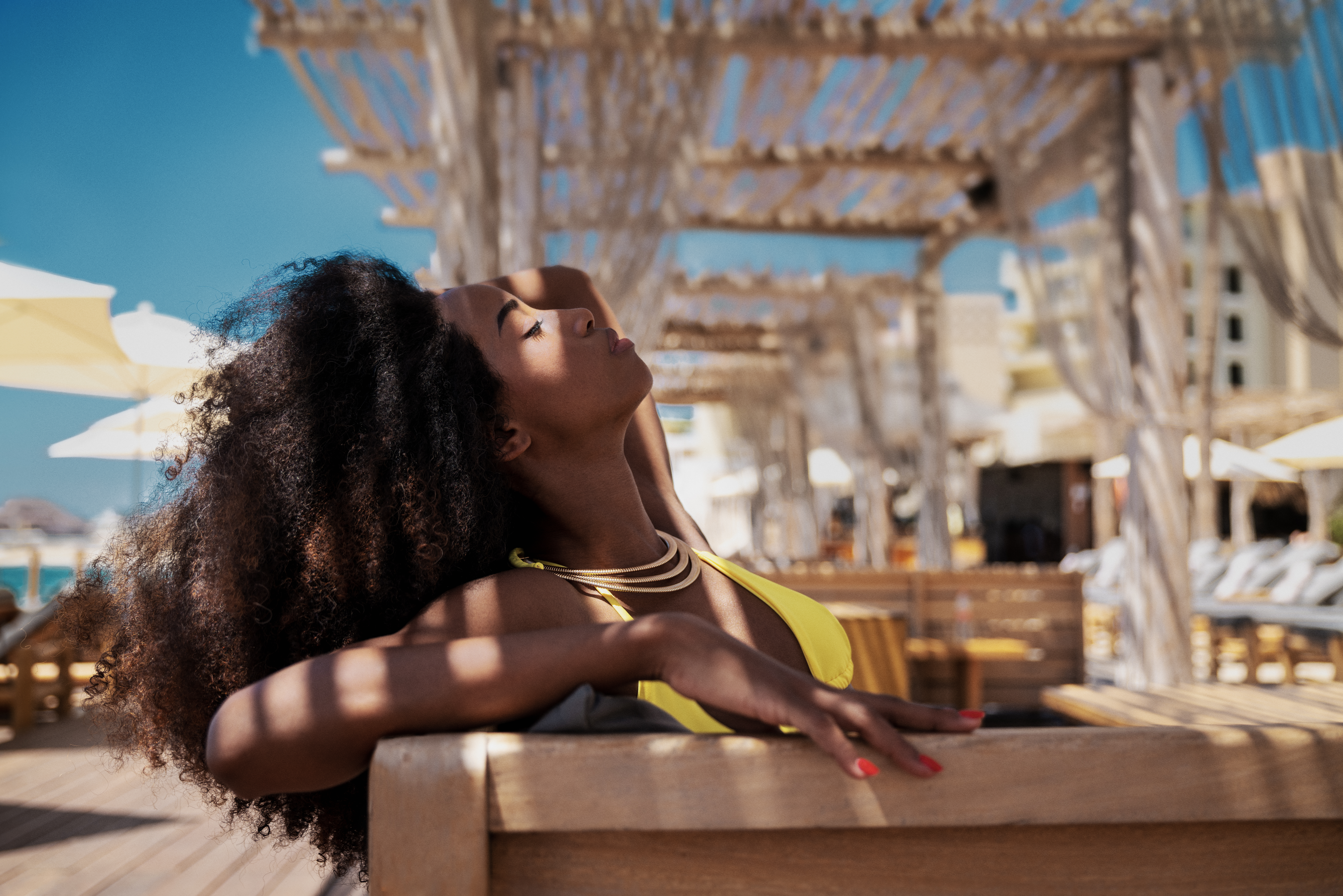 Curly-haired woman in yellow top, gold necklace, relaxing at sunny resort.