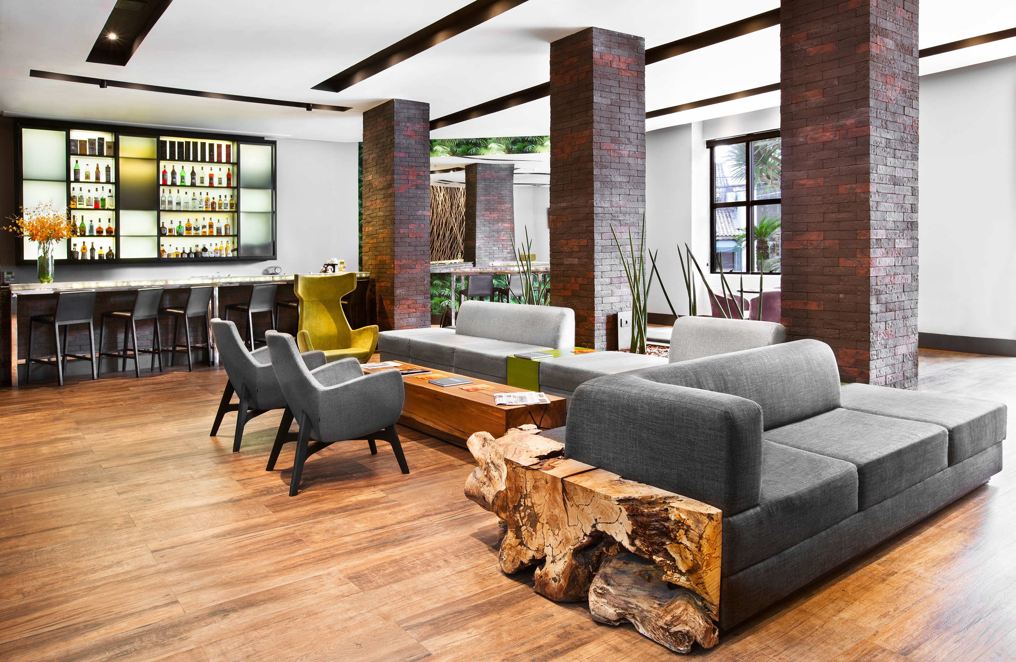 Modern hotel lobby lounge with bar, grey sofas, brick pillars, and wood flooring.