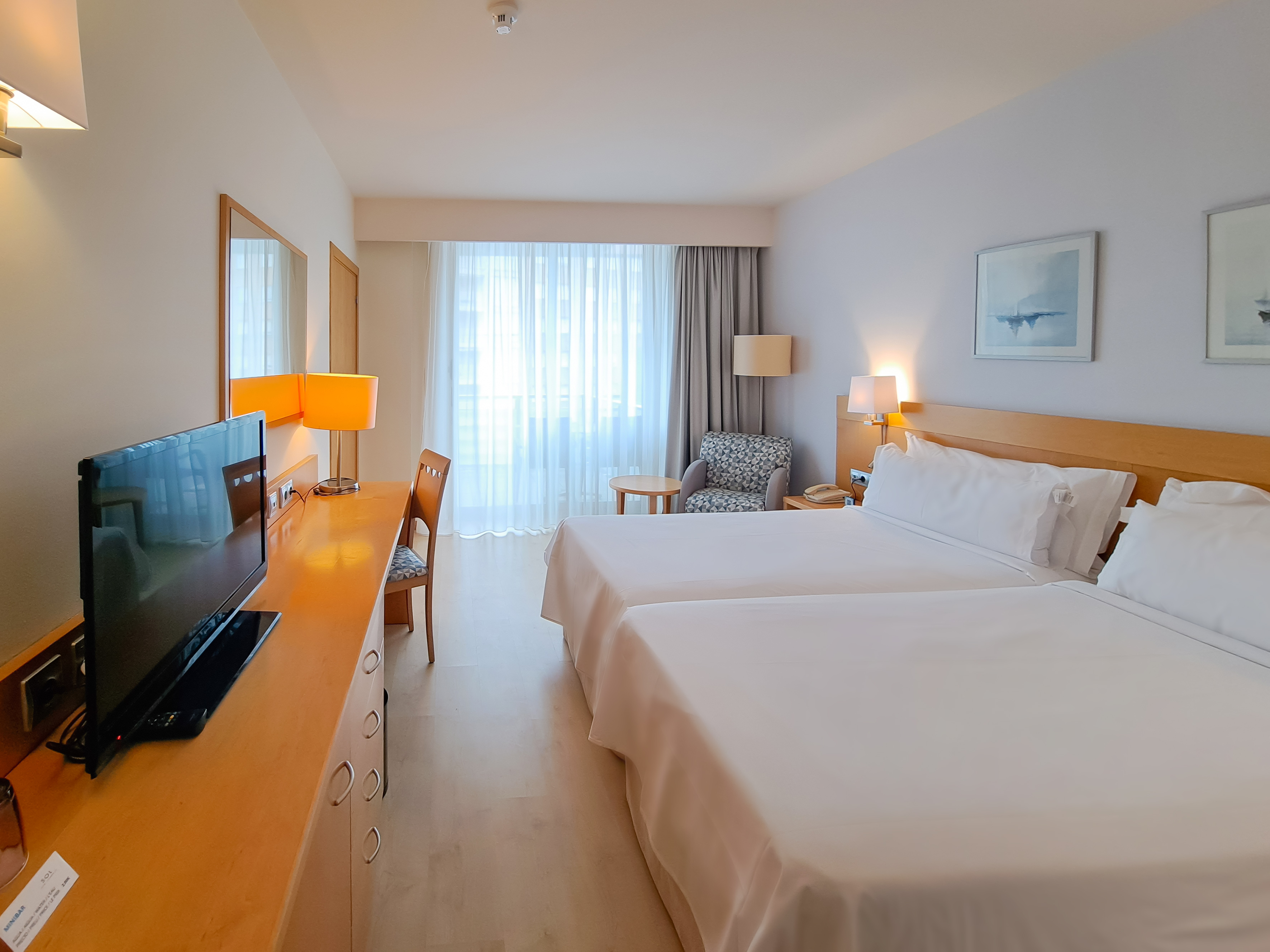 Bright hotel room with twin beds, wooden desk, TV, and armchair by window.