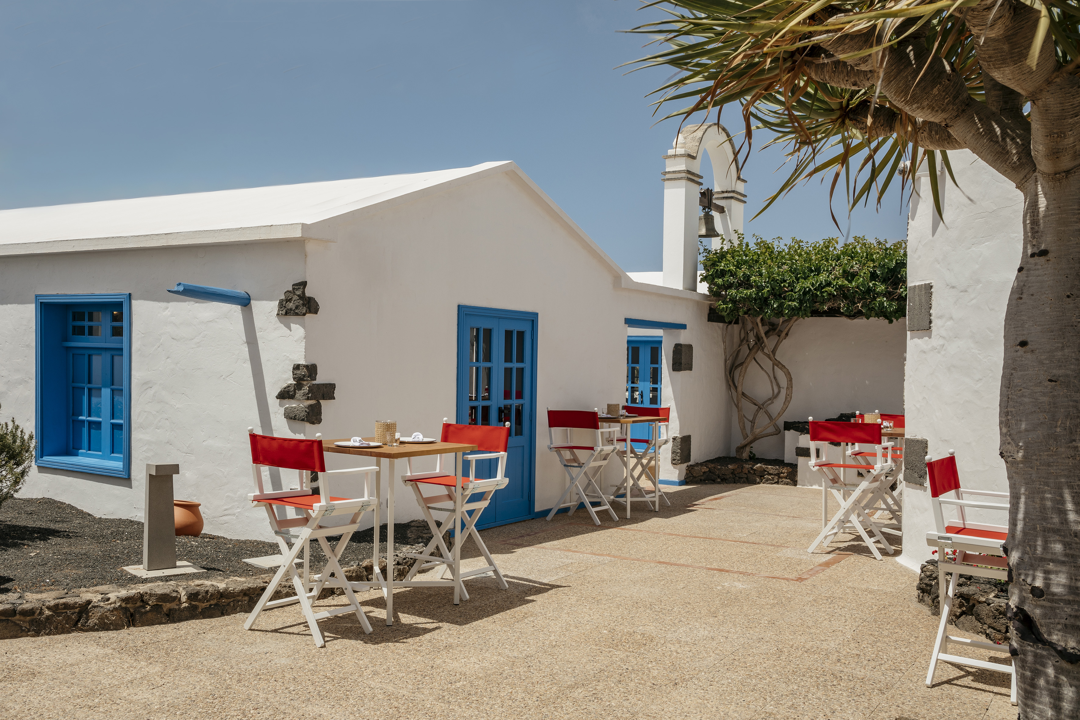 Sunny outdoor cafe patio with white architecture, blue windows, and red chairs.