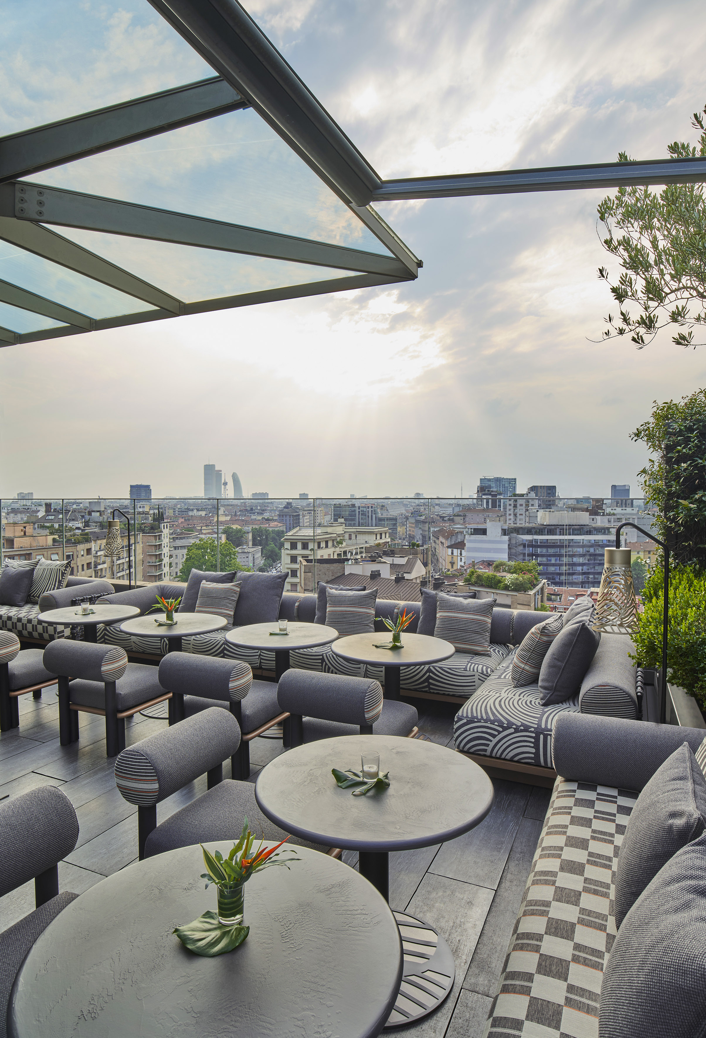Modern rooftop bar lounge with grey seating, glass canopy, and city view.
