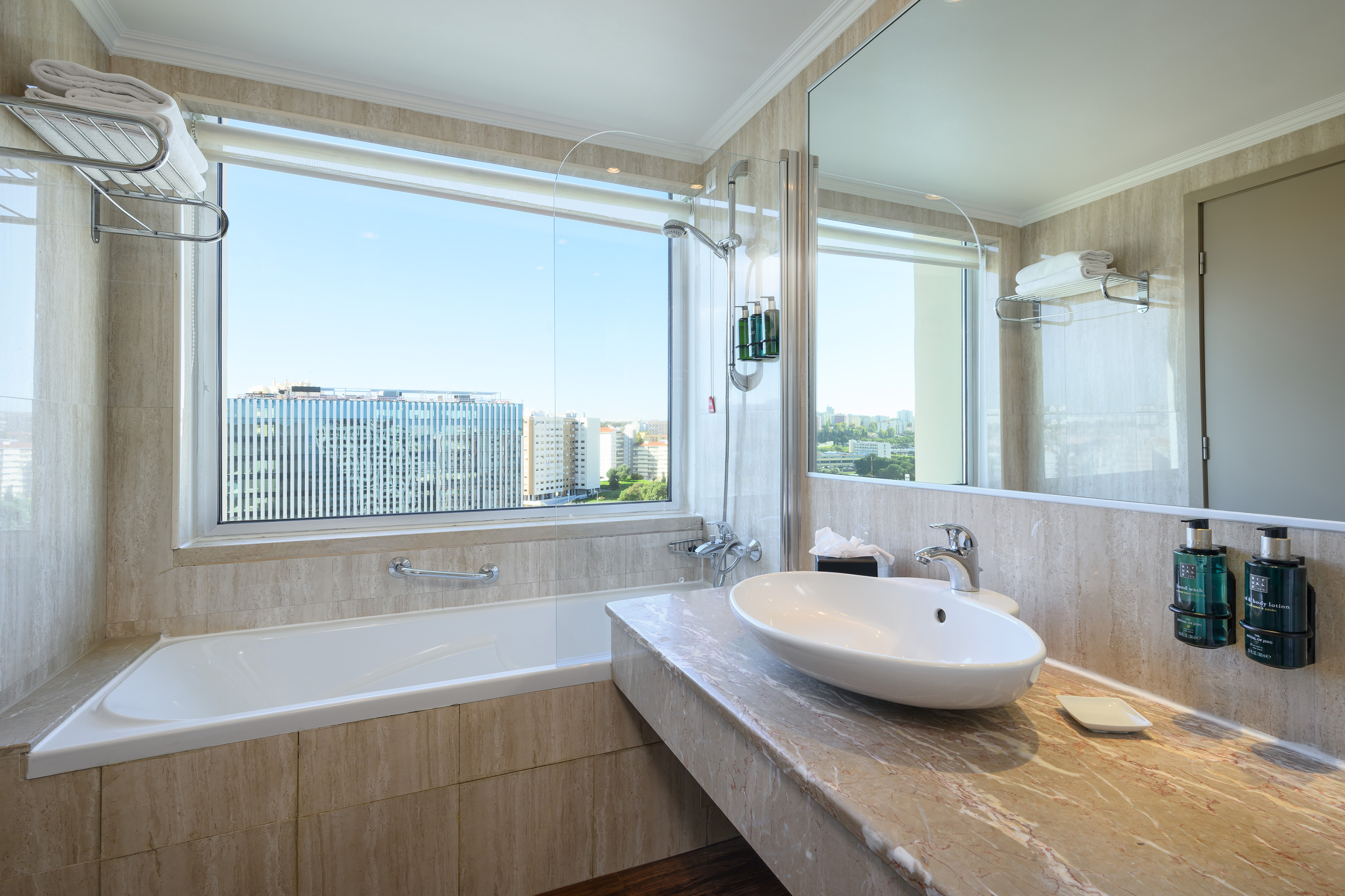 Modern hotel bathroom with city view window, bathtub, and marble vanity.