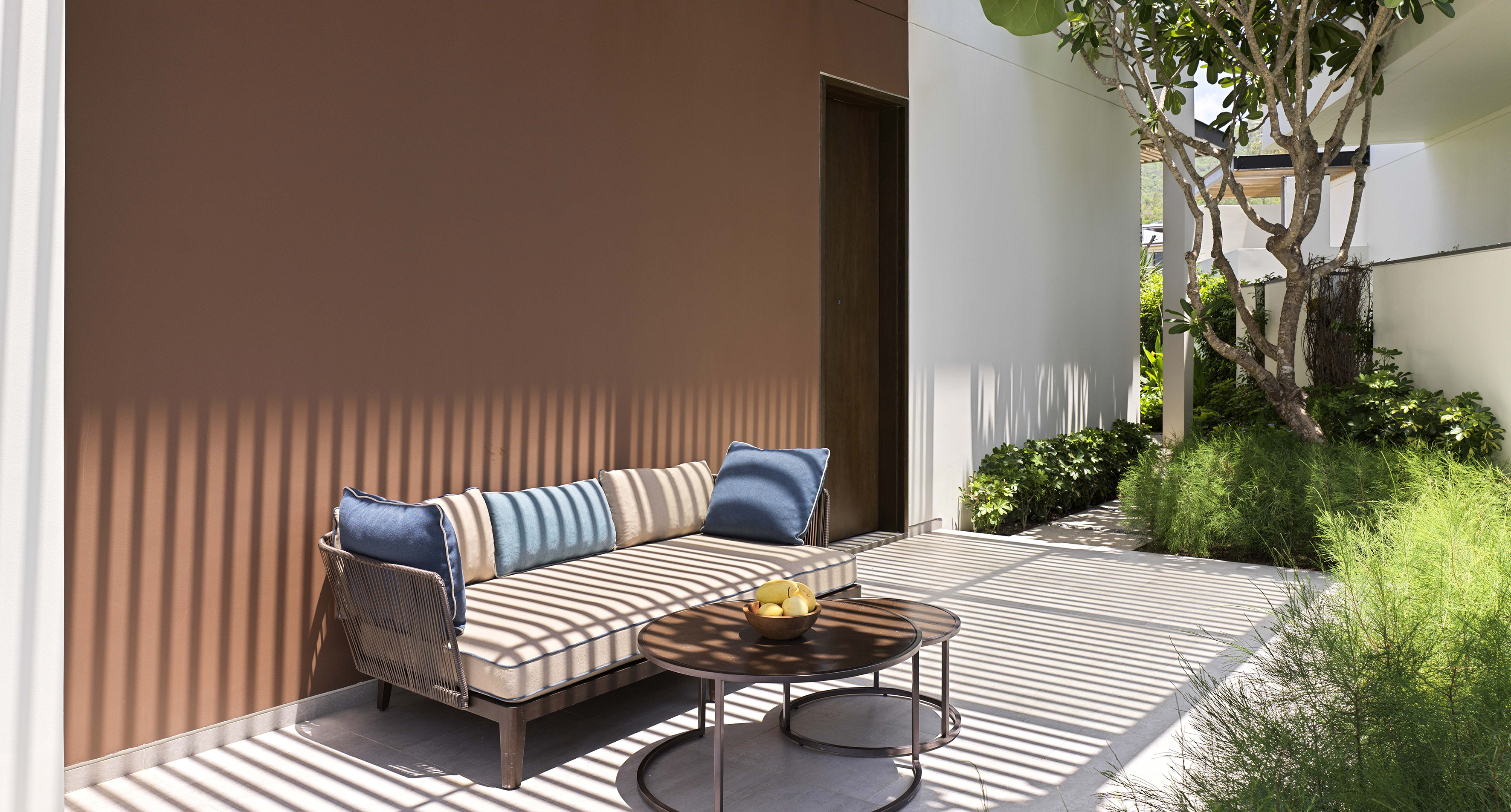 Sunny outdoor patio with modern daybed, coffee tables, and tropical garden.