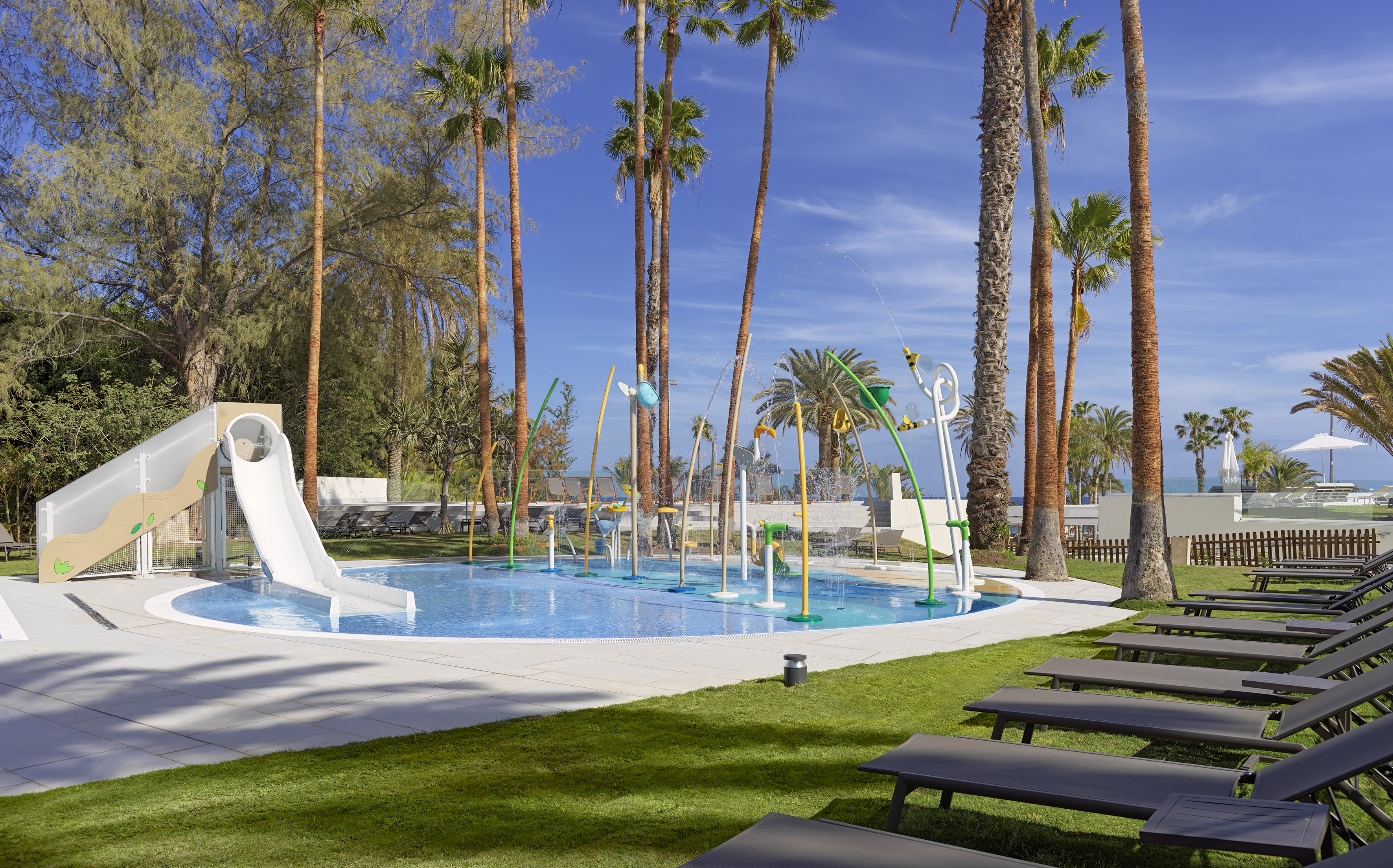 Kids' splash pad with water slide, palm trees, and lounge chairs at a sunny resort.