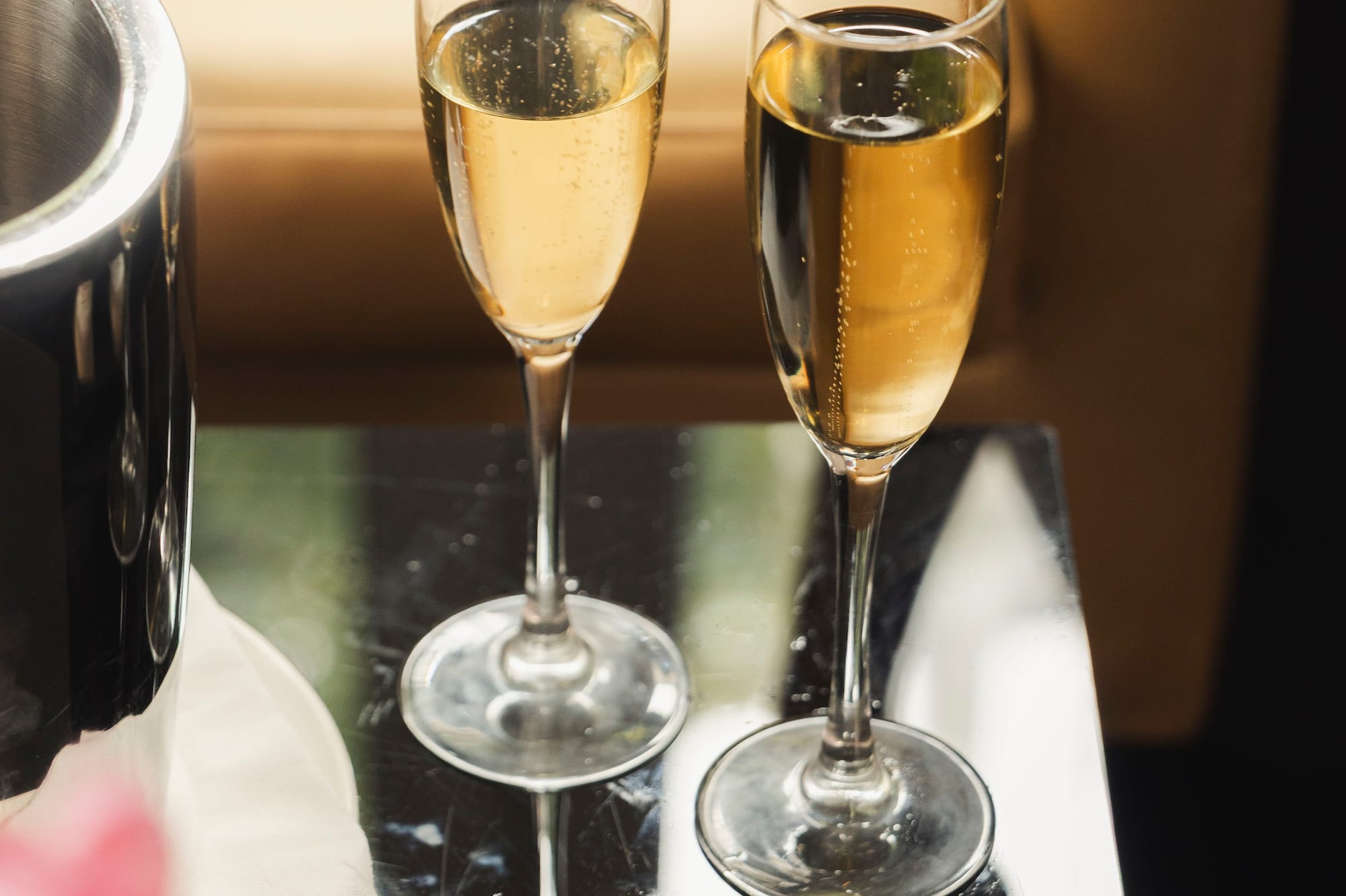 Two champagne flutes with sparkling wine, ice bucket, pink flowers on dark table.