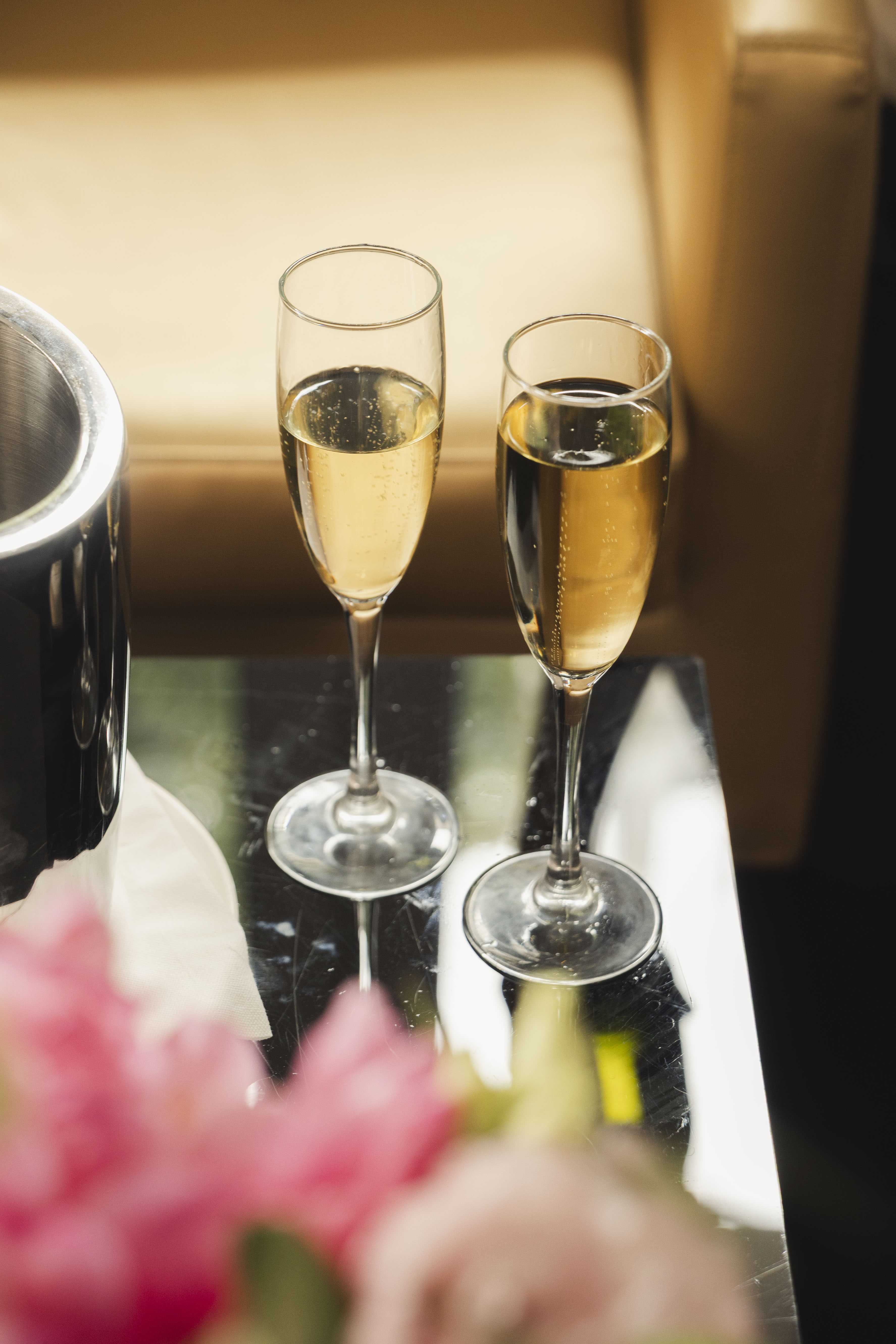 Two champagne flutes with sparkling wine, ice bucket, pink flowers on dark table.