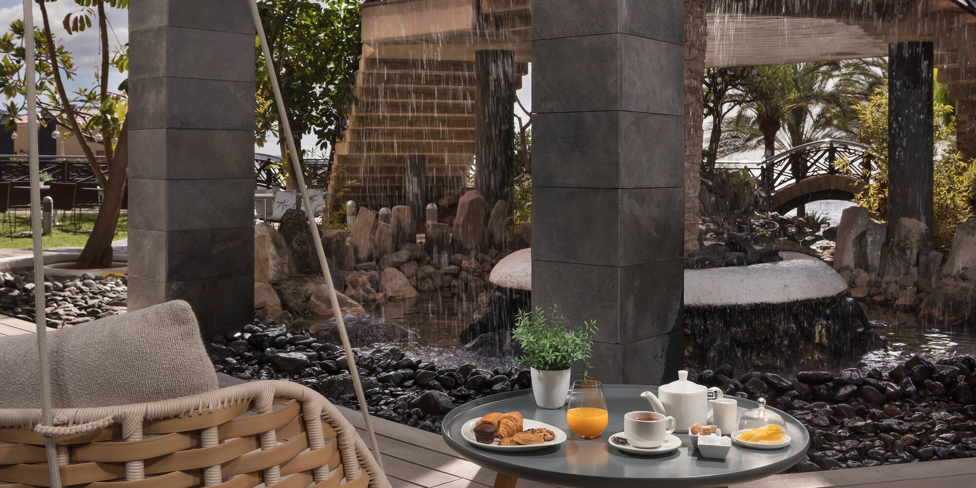Outdoor resort breakfast by waterfall: hanging chair, juice, tea, pastries.