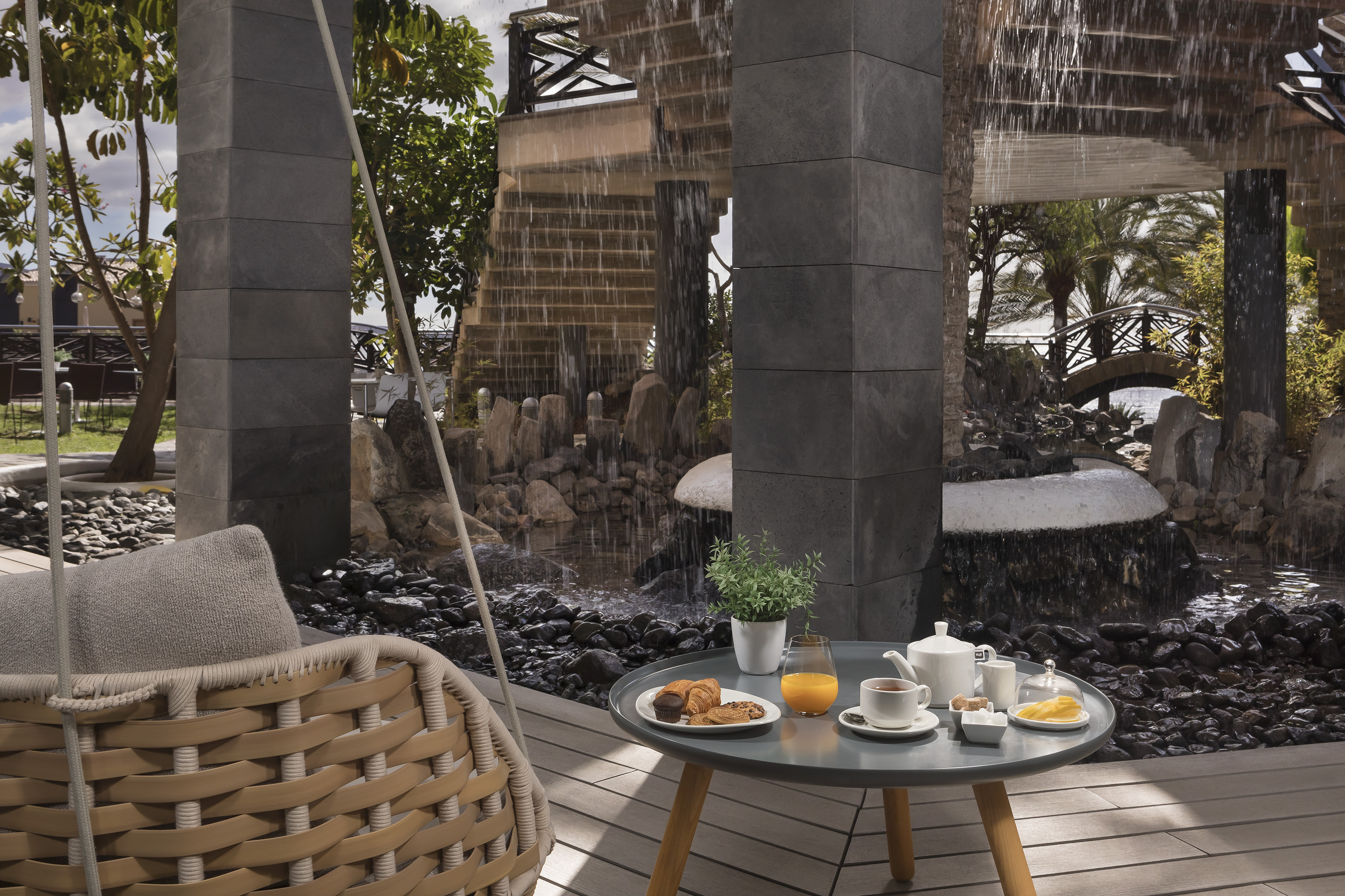 Outdoor resort breakfast by waterfall: hanging chair, juice, tea, pastries.