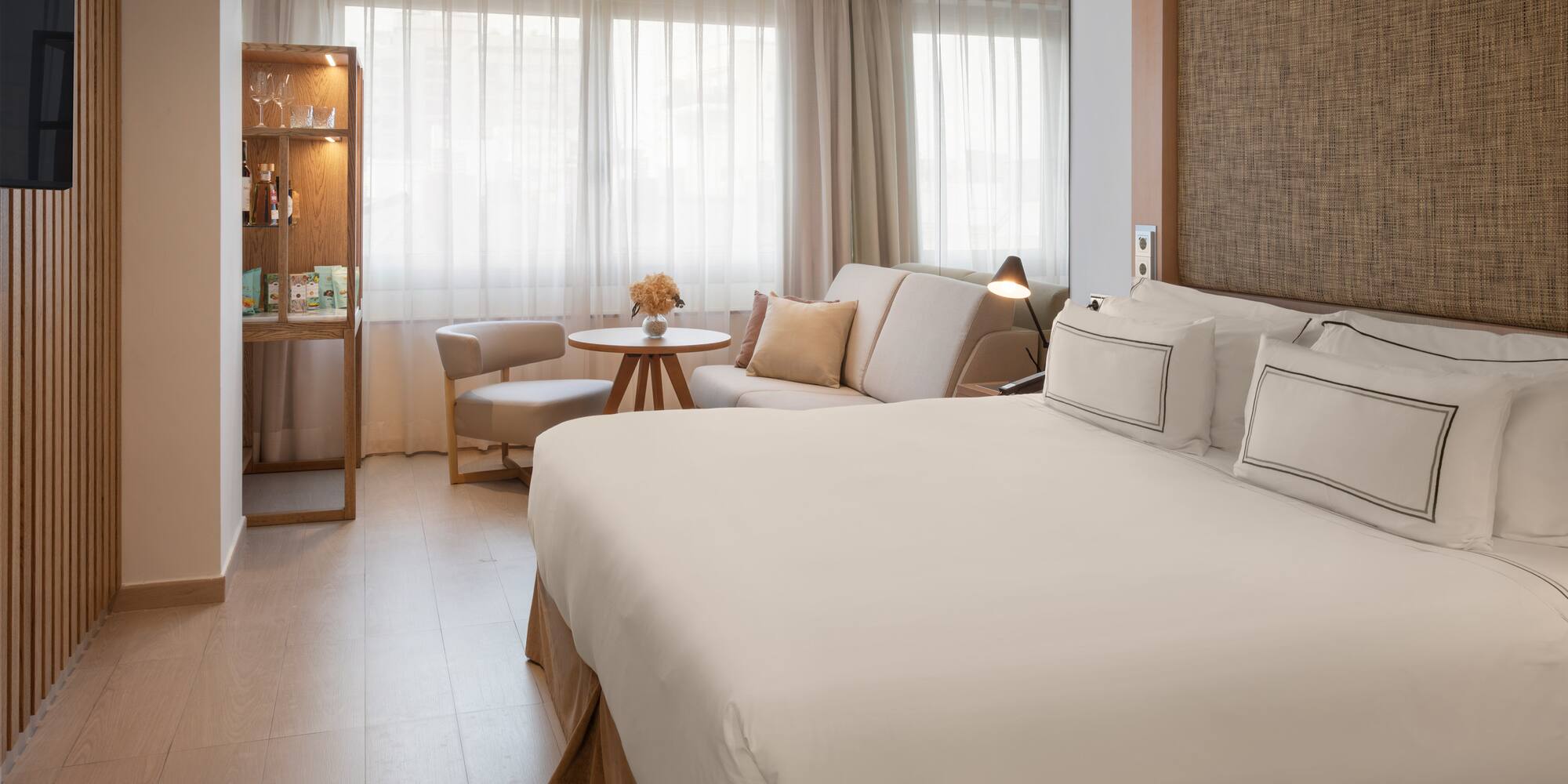 Bright hotel room with a large bed, sofa, and light wooden accents.