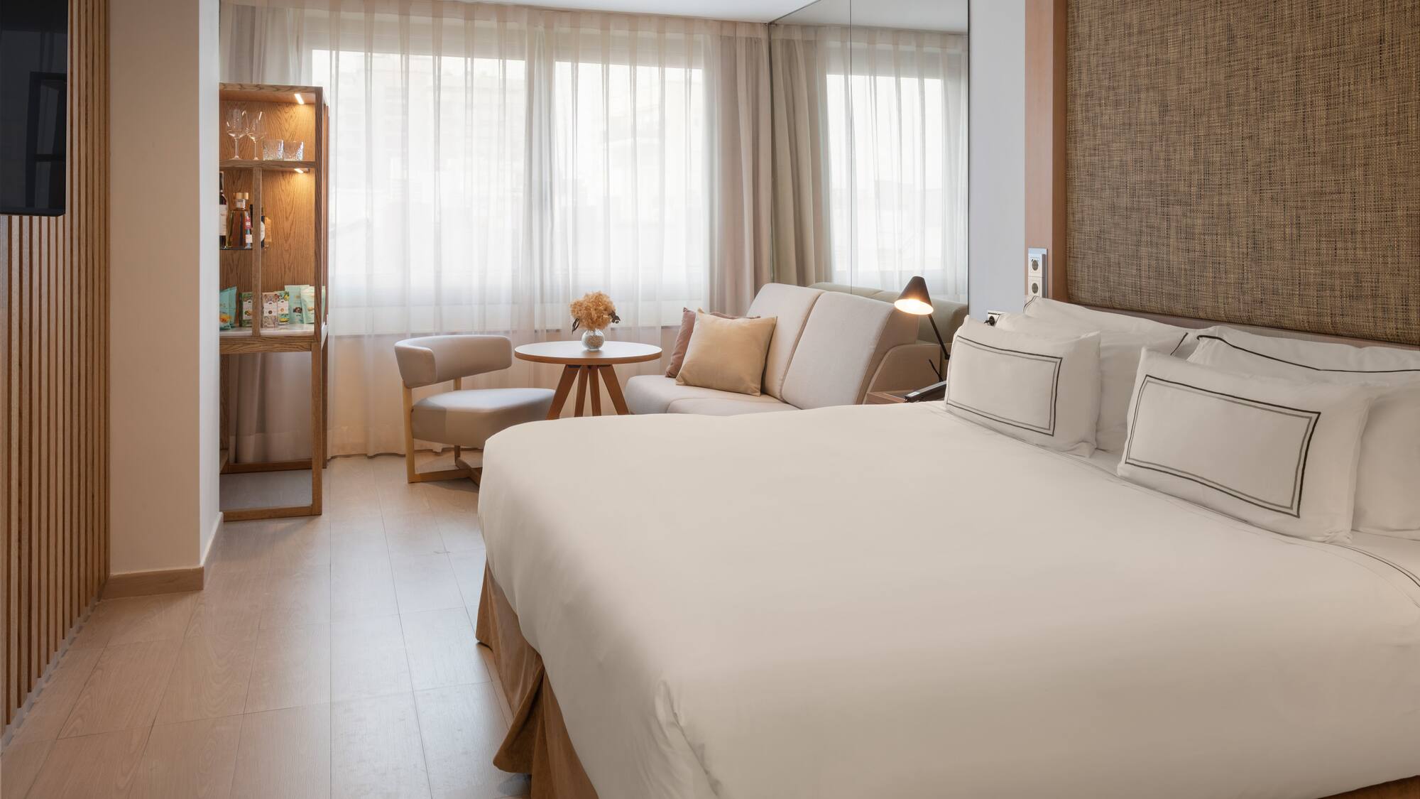 Bright hotel room with a large bed, sofa, and light wooden accents.