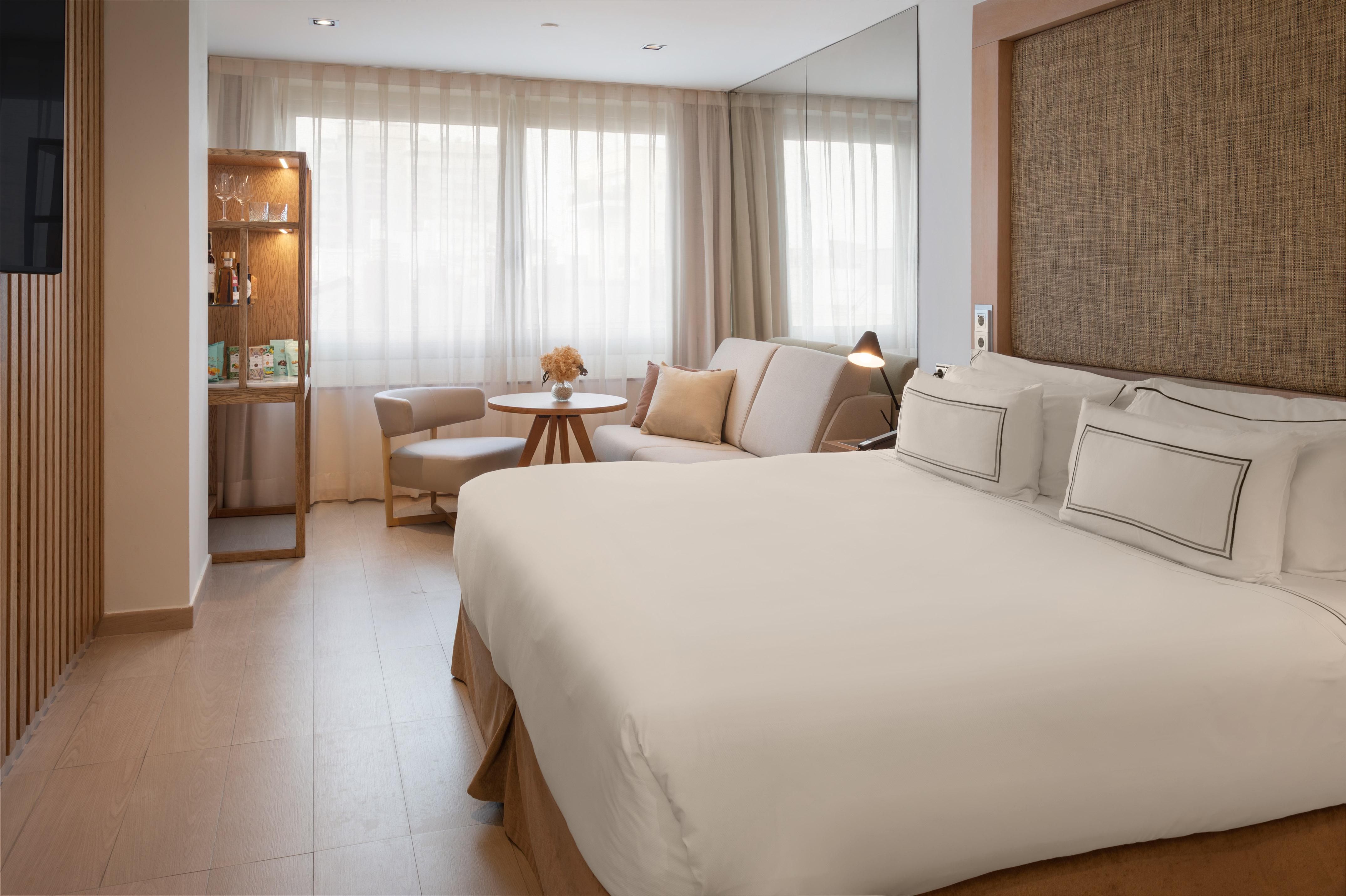 Habitación de hotel moderna con cama doble, sofá, minibar y ventanales.