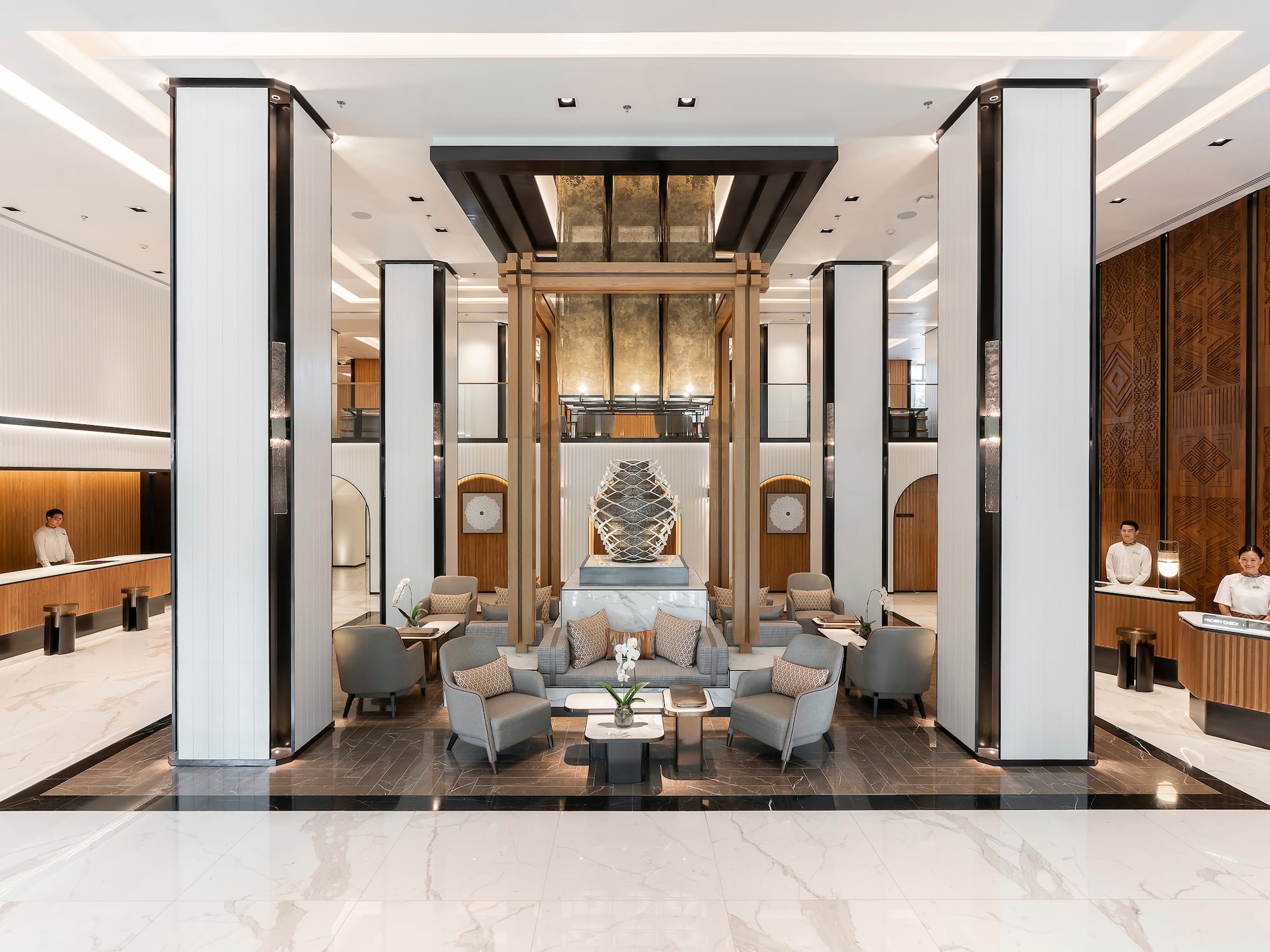 Modern hotel lobby with reception desks, central lounge seating, and art sculpture.