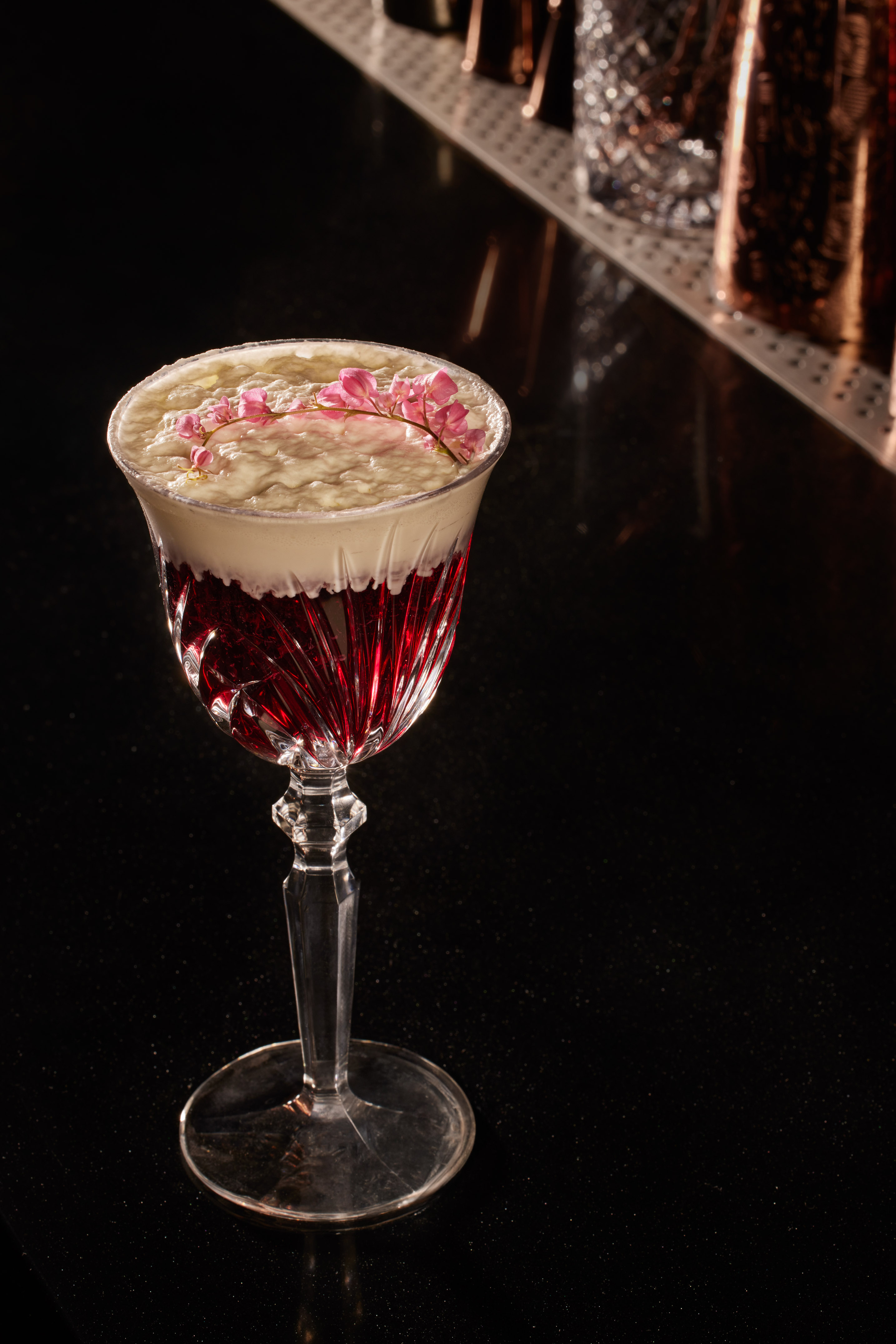 Elegant red cocktail with foamy top, pink floral garnish, on a bar.