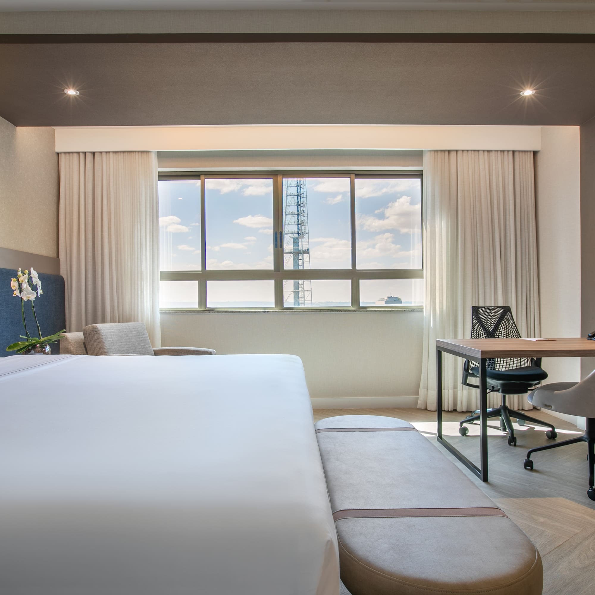 Contemporary hotel bedroom with white bed, desk area, and city skyline view.