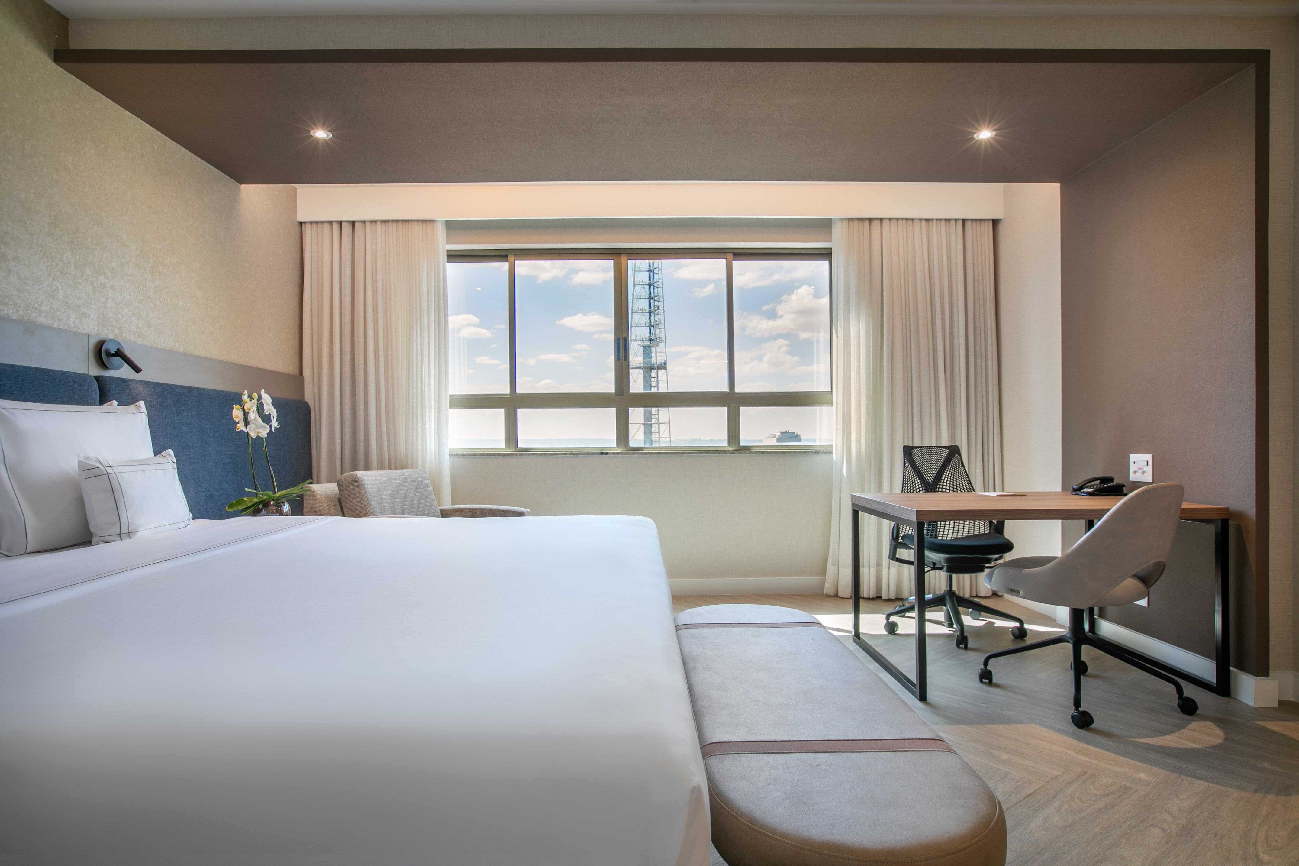 Contemporary hotel bedroom with white bed, desk area, and city skyline view.
