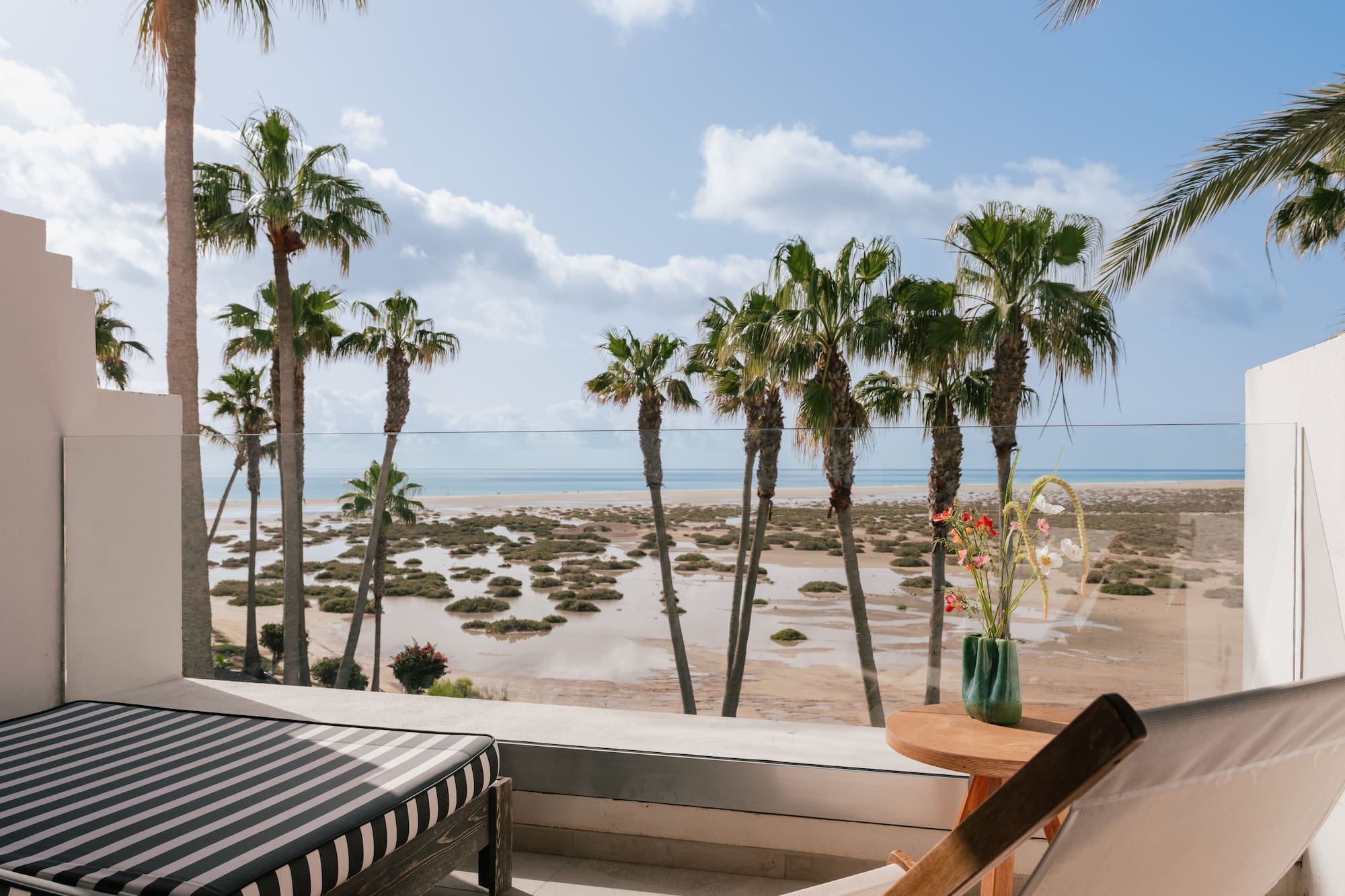 Terraza con vistas al mar, palmeras, playa arenosa y tumbonas.