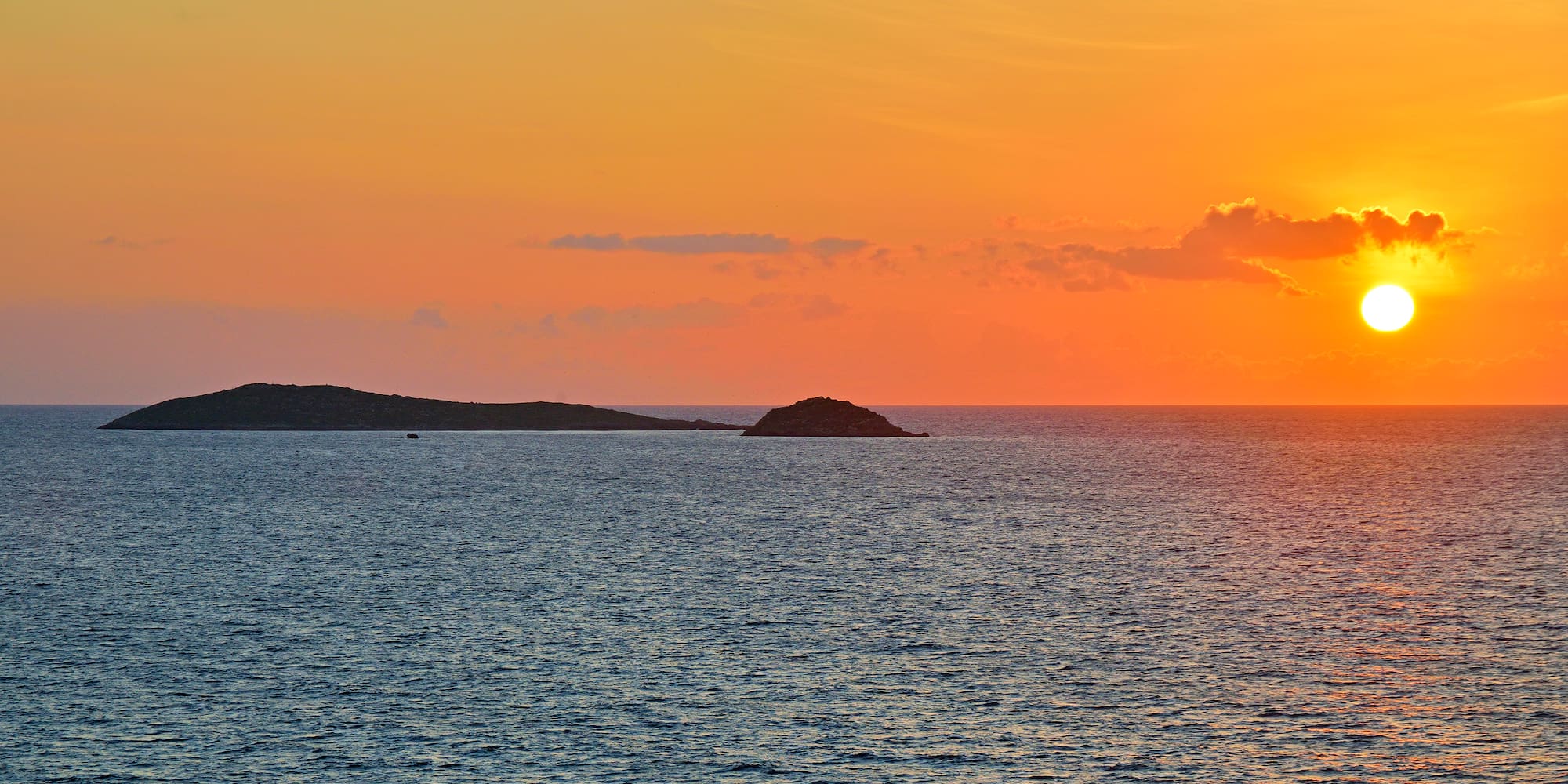Sunset seascape with vibrant orange sky, islands, and bright sun.
