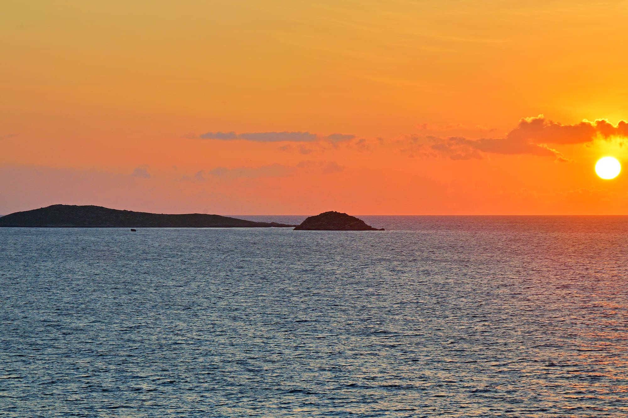 Orange sunset over ocean with two small islands and bright sun.