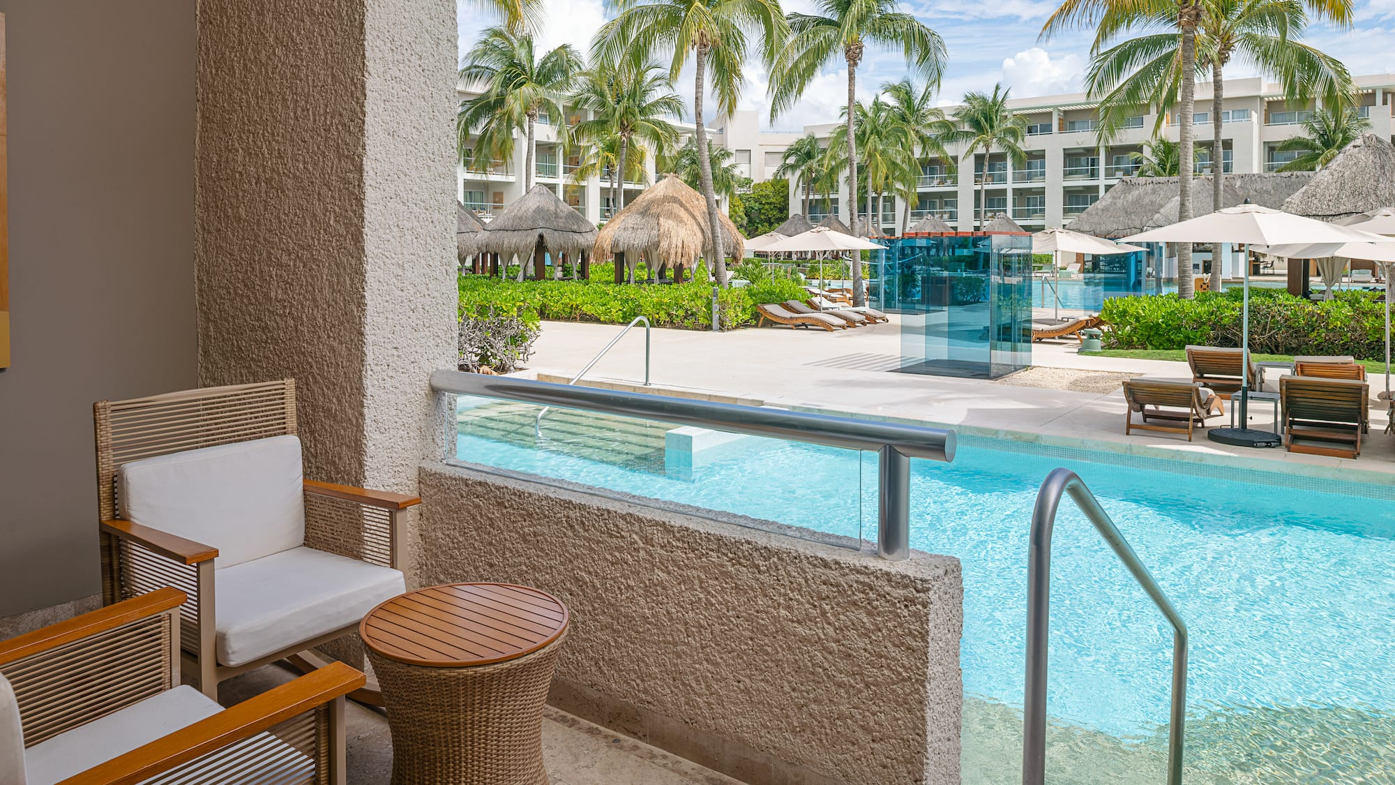 Patio with chairs overlooking a tropical resort pool, palm trees, and hotel buildings.