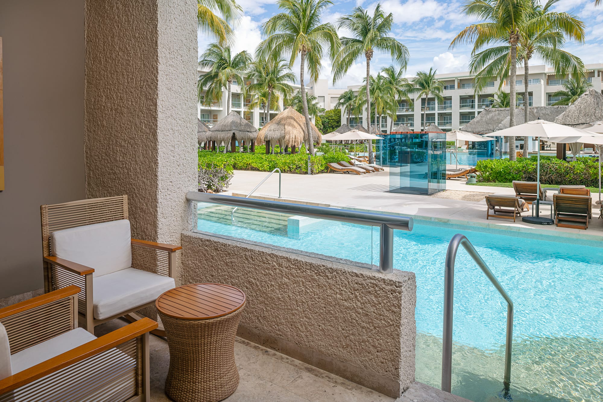 Patio with chairs overlooking a tropical resort pool, palm trees, and hotel buildings.