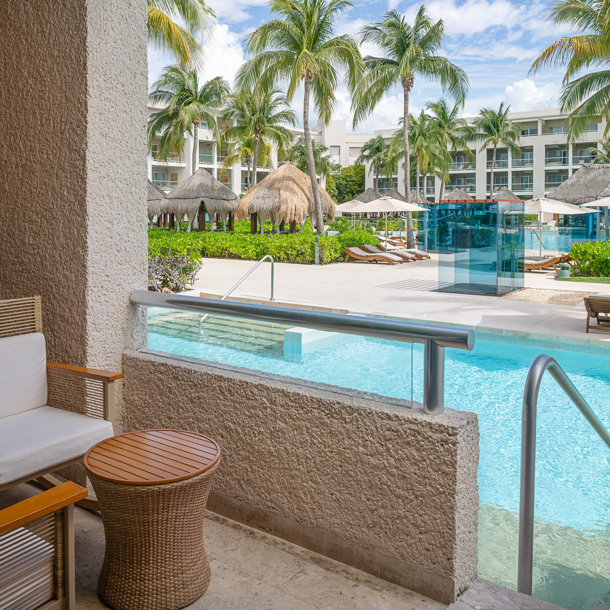 Patio with chairs overlooking a tropical resort pool, palm trees, and hotel buildings.