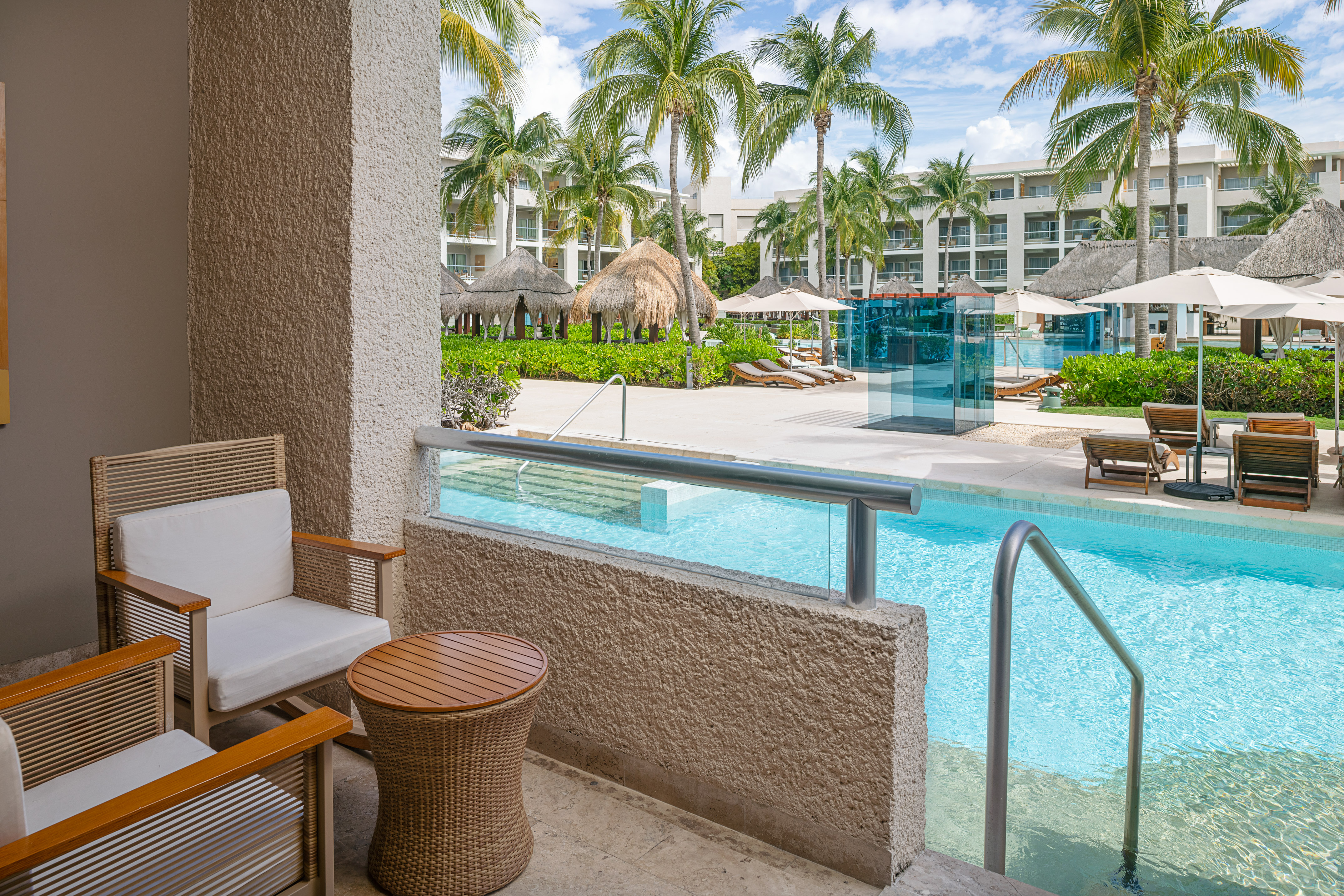 Patio with chairs overlooking a tropical resort pool, palm trees, and hotel buildings.