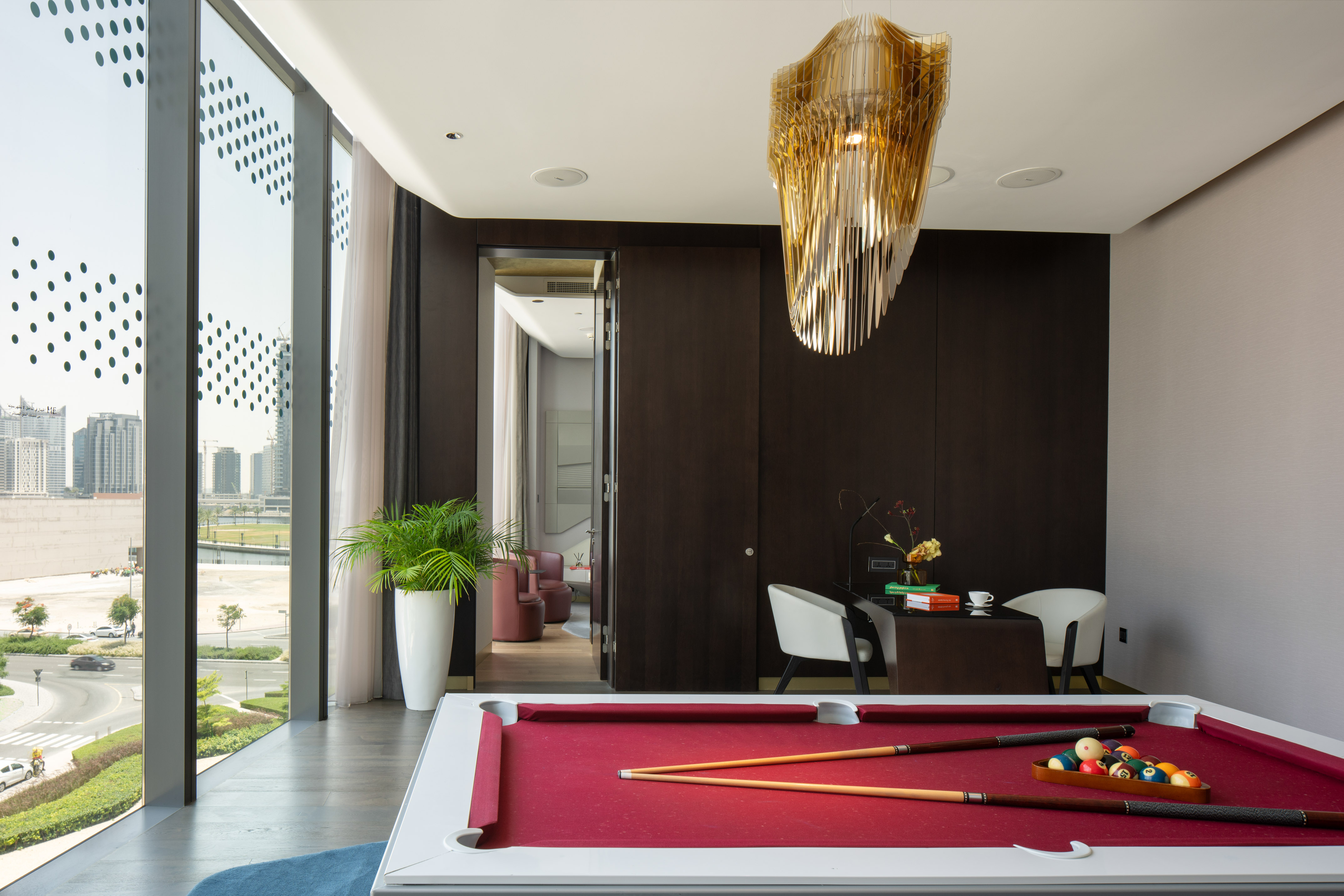 Modern game room with red pool table, gold chandelier, and city skyline view.