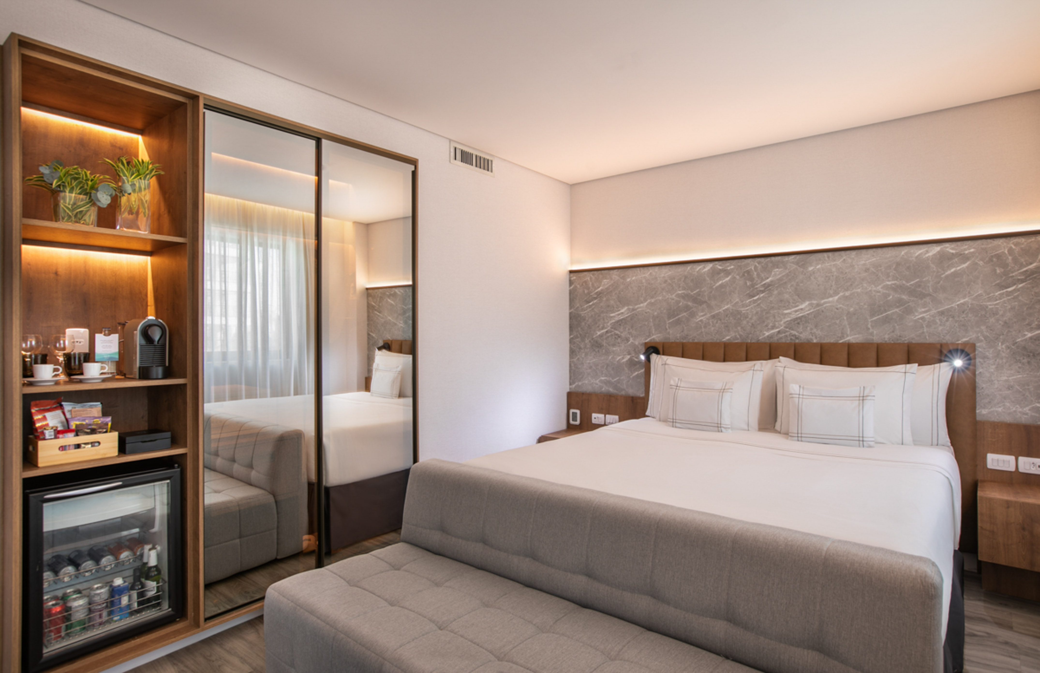 Modern hotel room: king bed, marble accent wall, mini-bar, and wooden shelves.