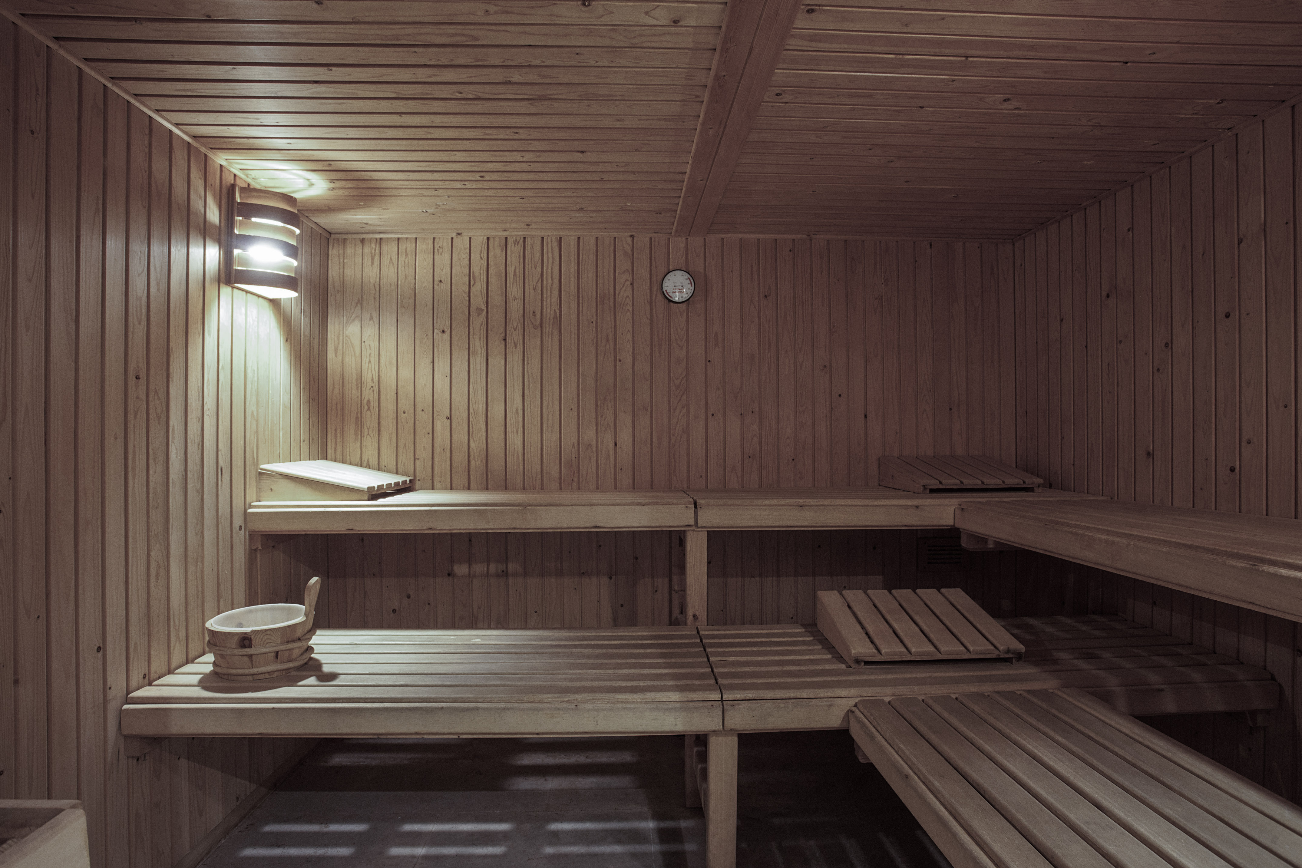 Wooden sauna room with multi-level benches, wall light, and bucket.