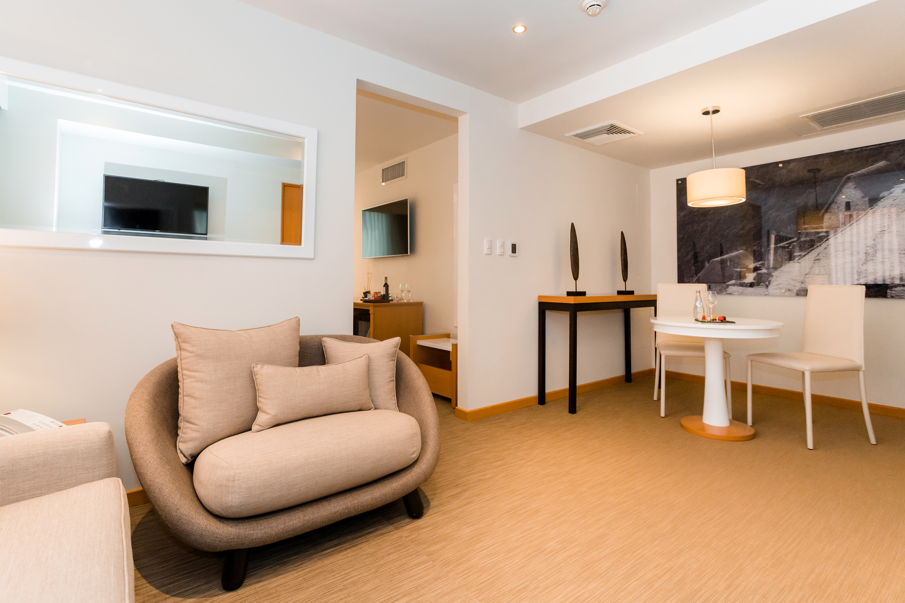 Modern hotel suite with living area, dining table, armchair, and neutral tones.