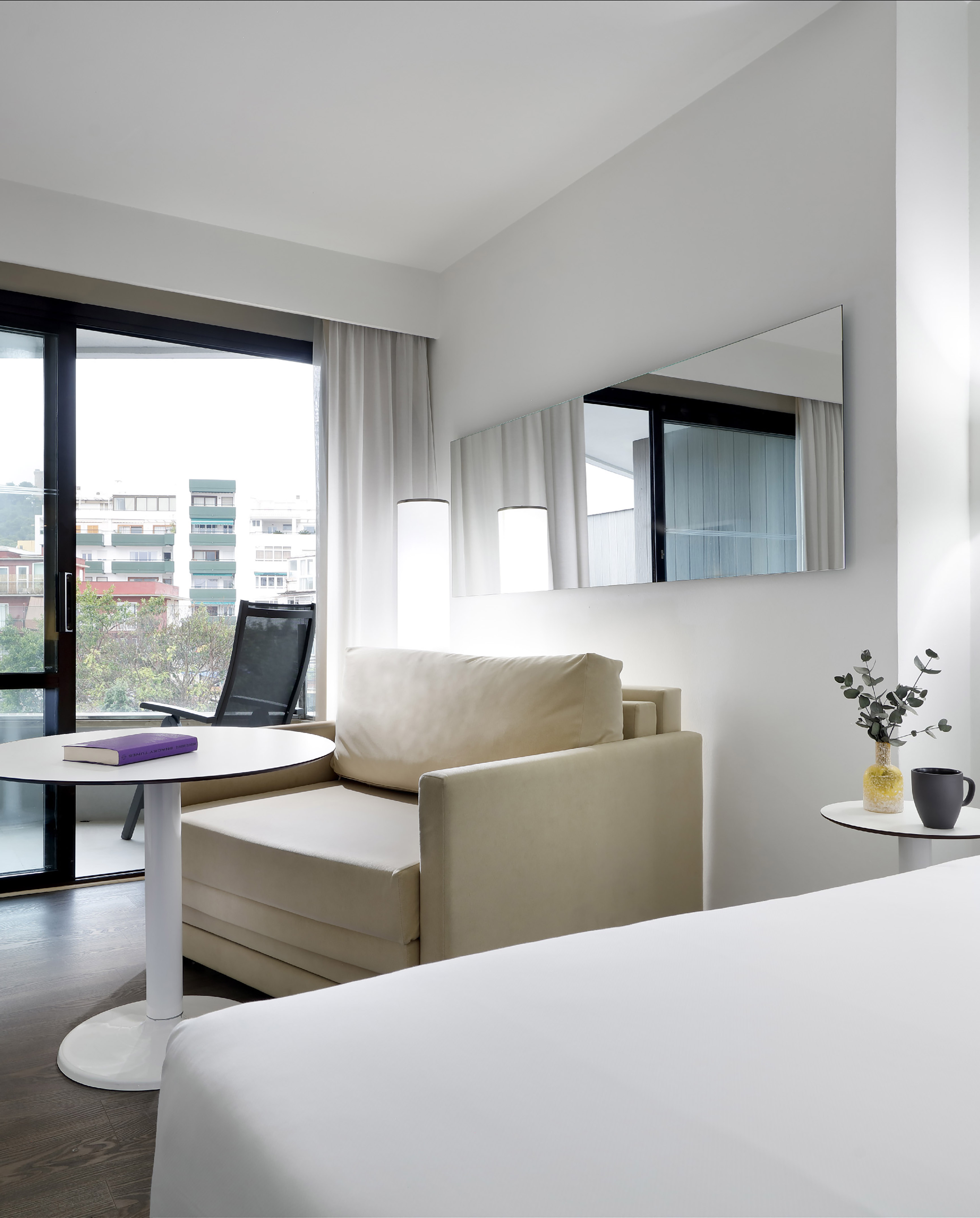 Bright modern hotel room with sofa, bed, city view balcony, and sleek mirror.