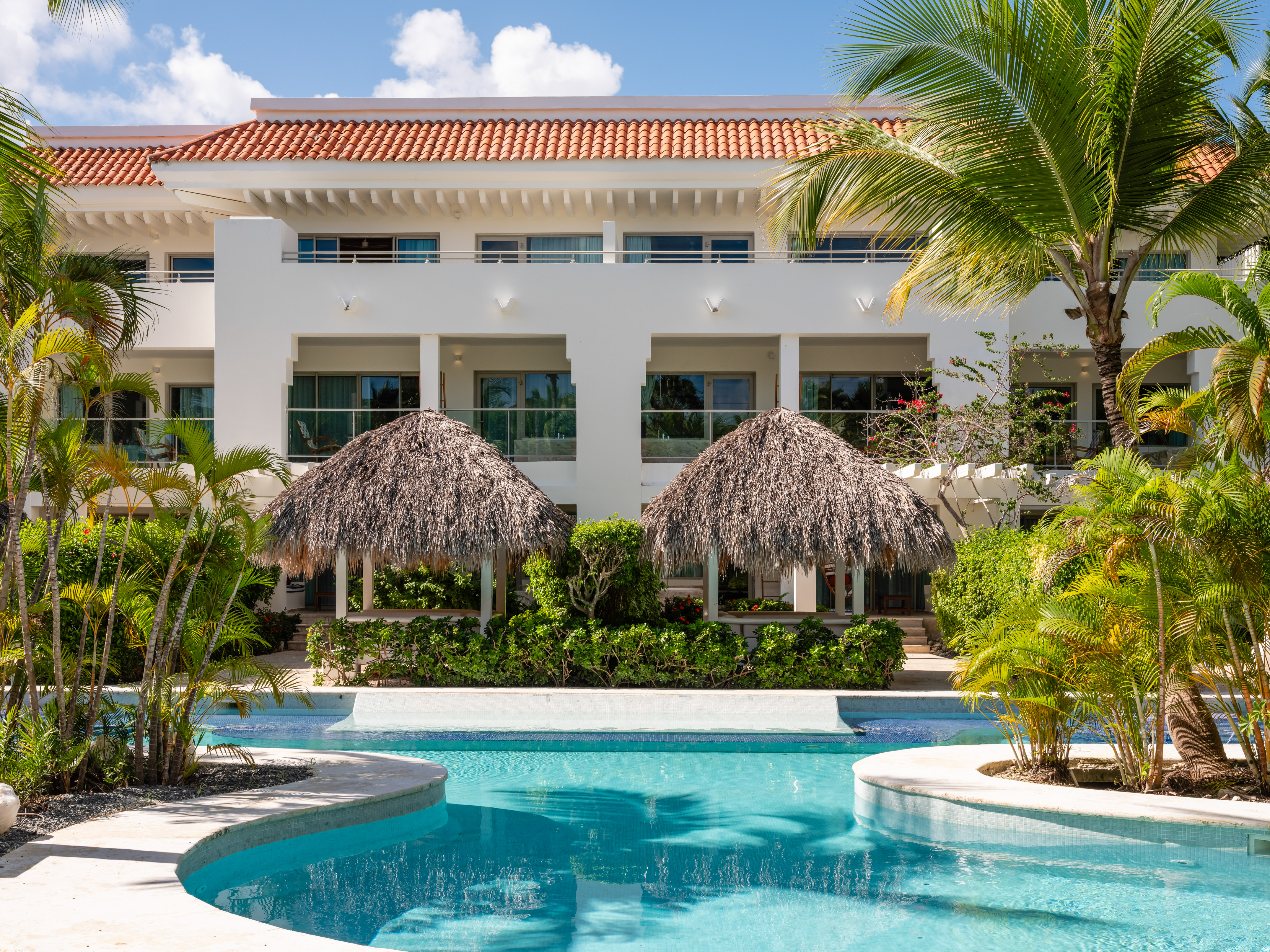 Tropical resort building with swimming pool, palm trees, and thatched gazebos.