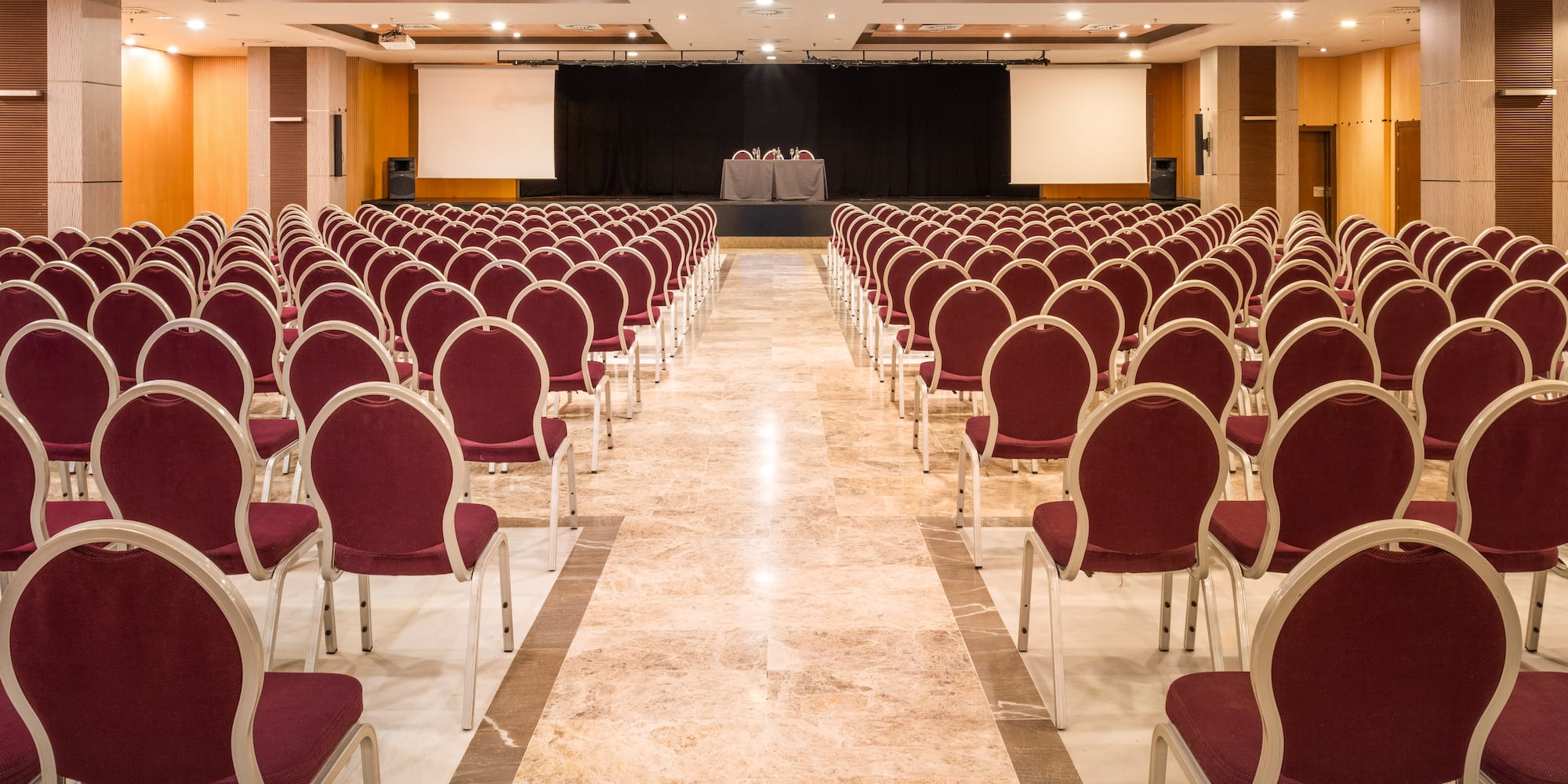 Empty conference room with red chairs, stage, and dual projector screens.
