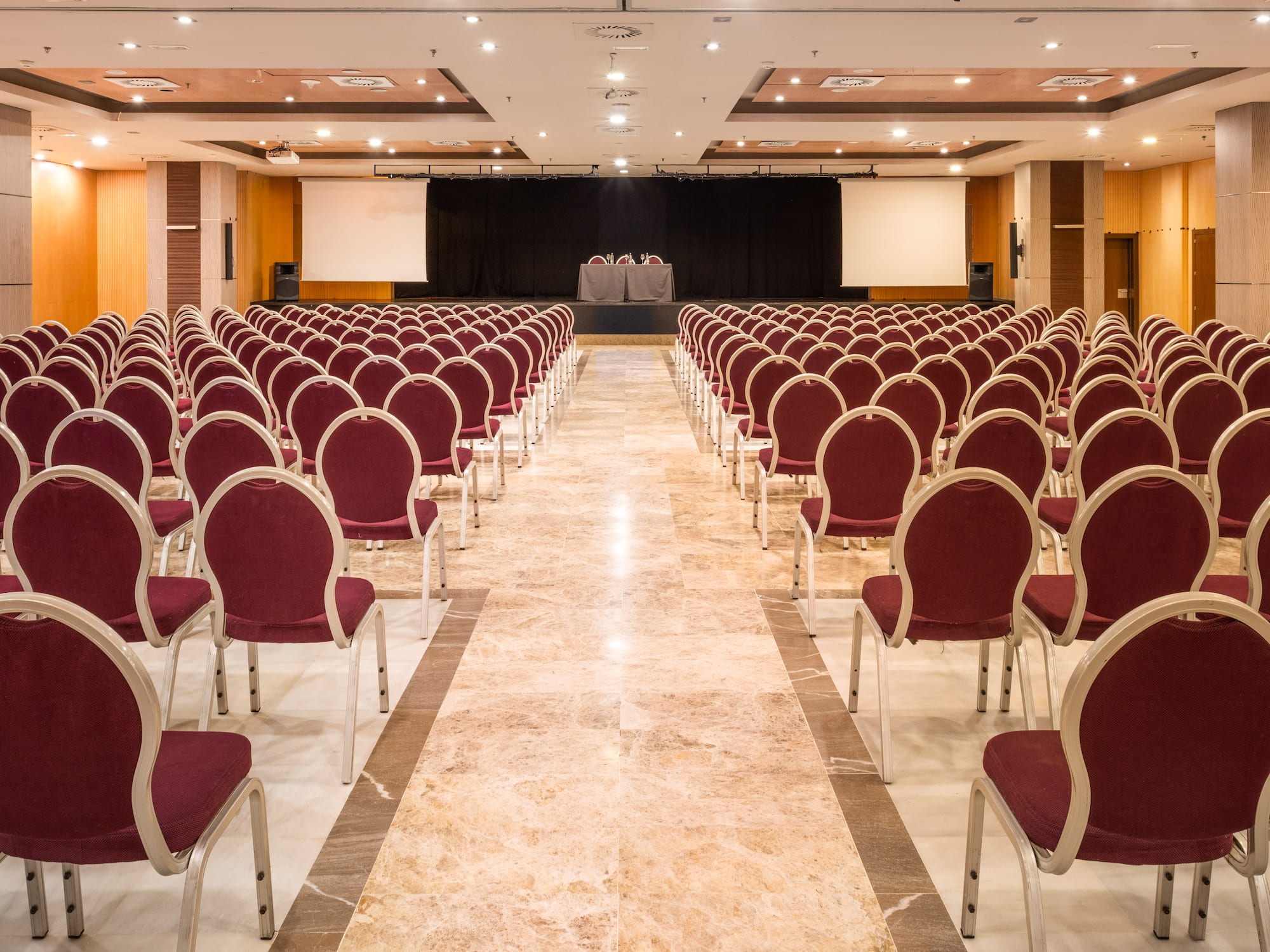 Large empty conference room with rows of red chairs, stage, and projector screens.