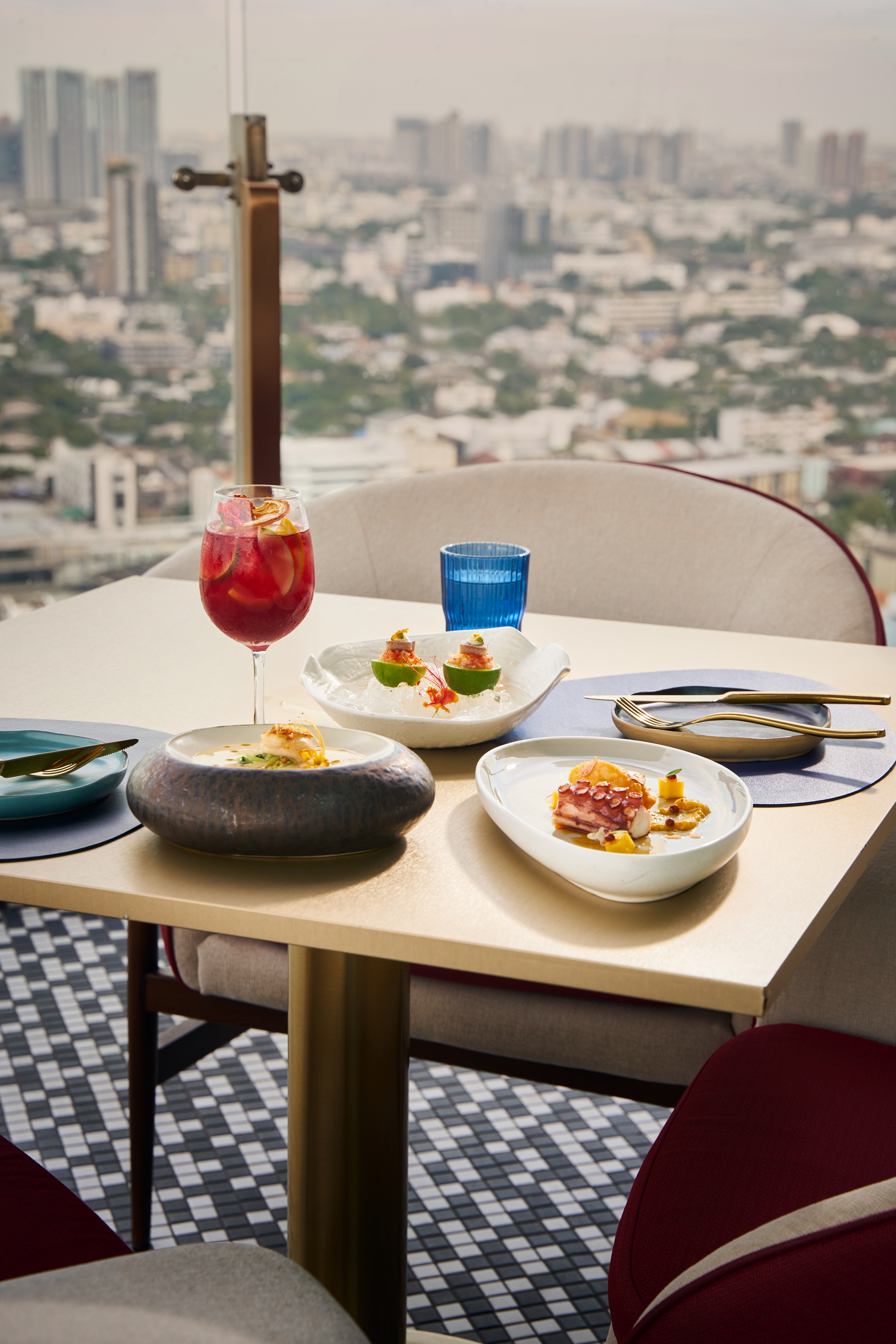 Gourmet dishes, red cocktail, and city skyline view from high-rise restaurant.