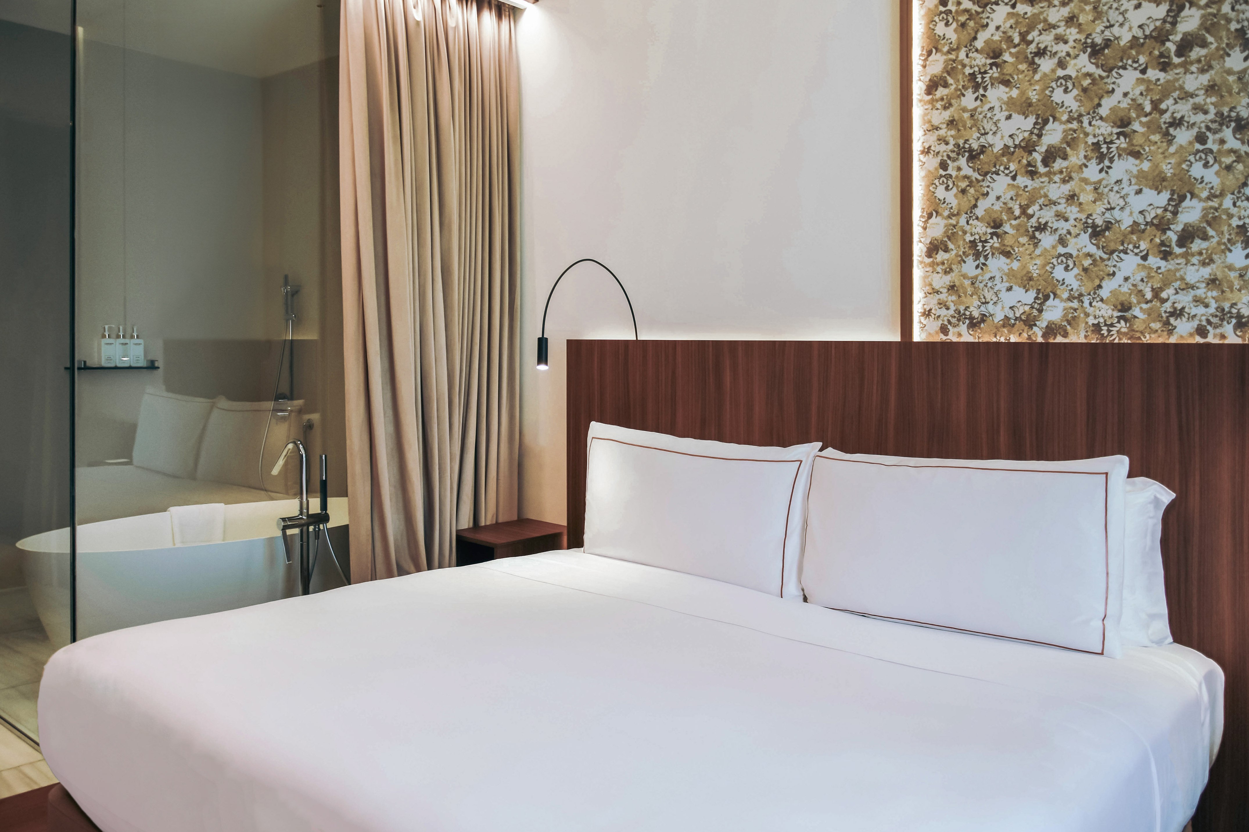 Stylish hotel room: white bed, wood headboard, bathtub visible through glass.