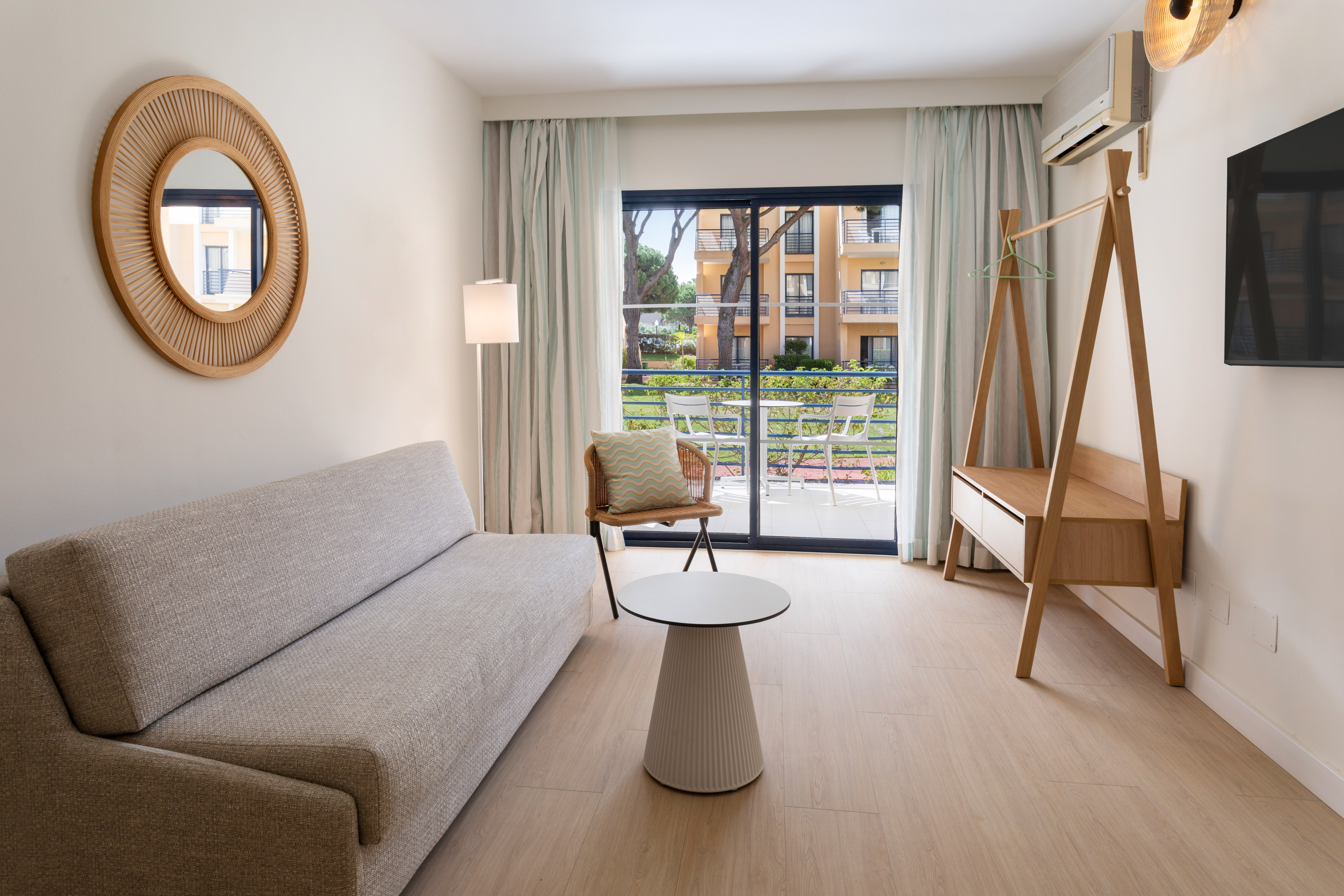 Bright modern hotel room with sofa, round mirror, and balcony overlooking resort.