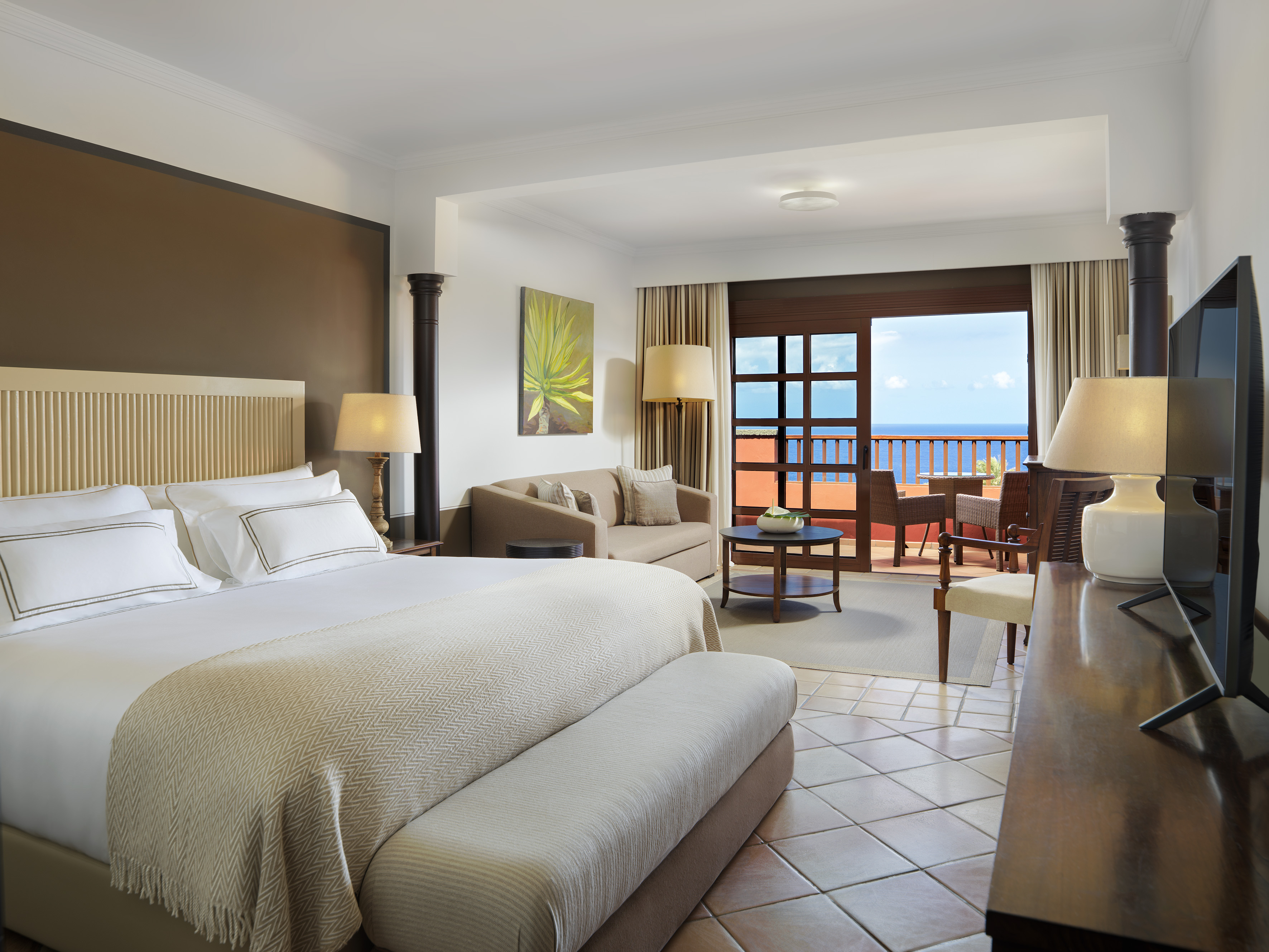 Hotel suite with king bed, seating area, and ocean view balcony.