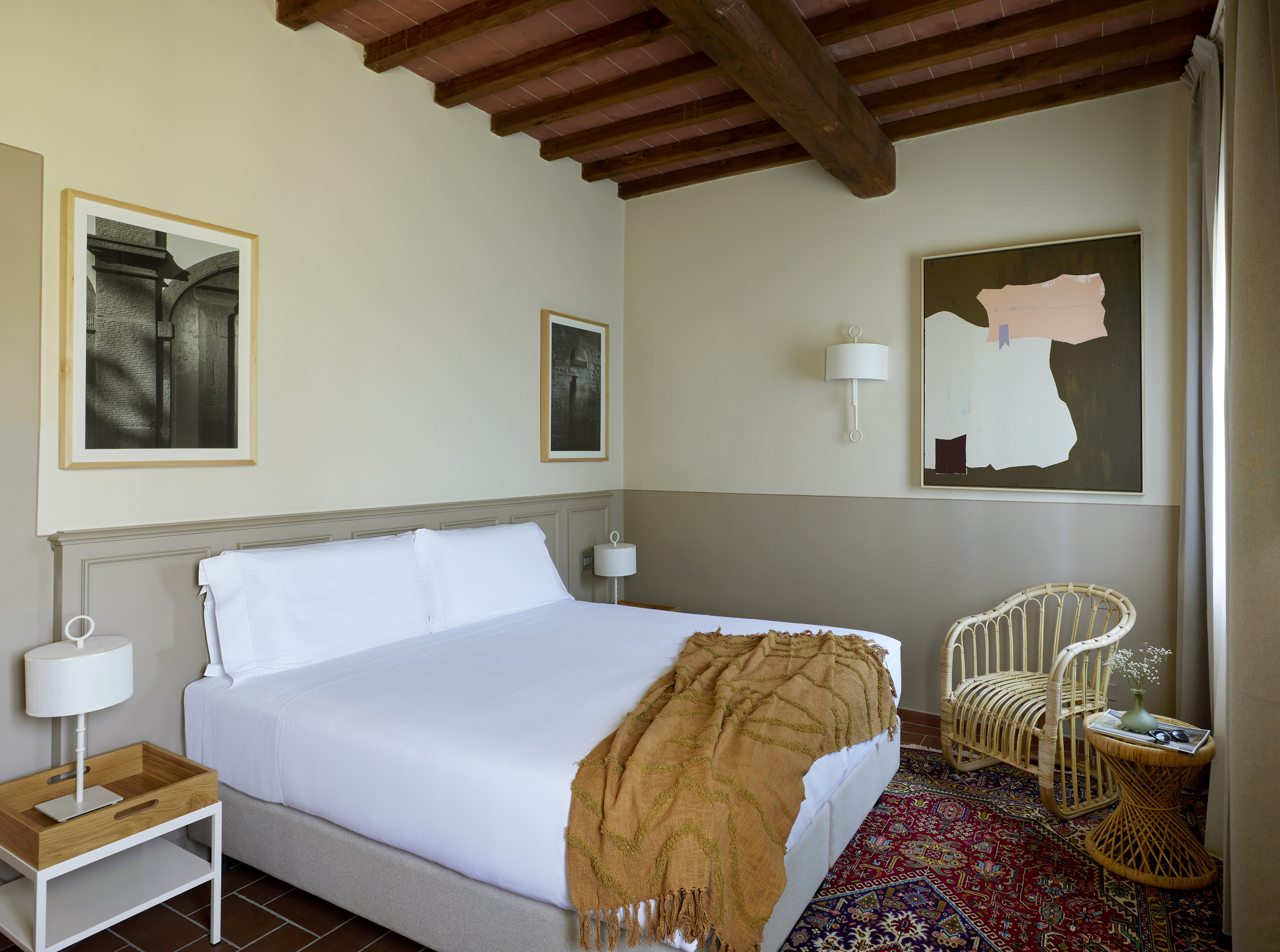 Rustic modern bedroom with beamed ceiling, white bed, terracotta floor, and art.