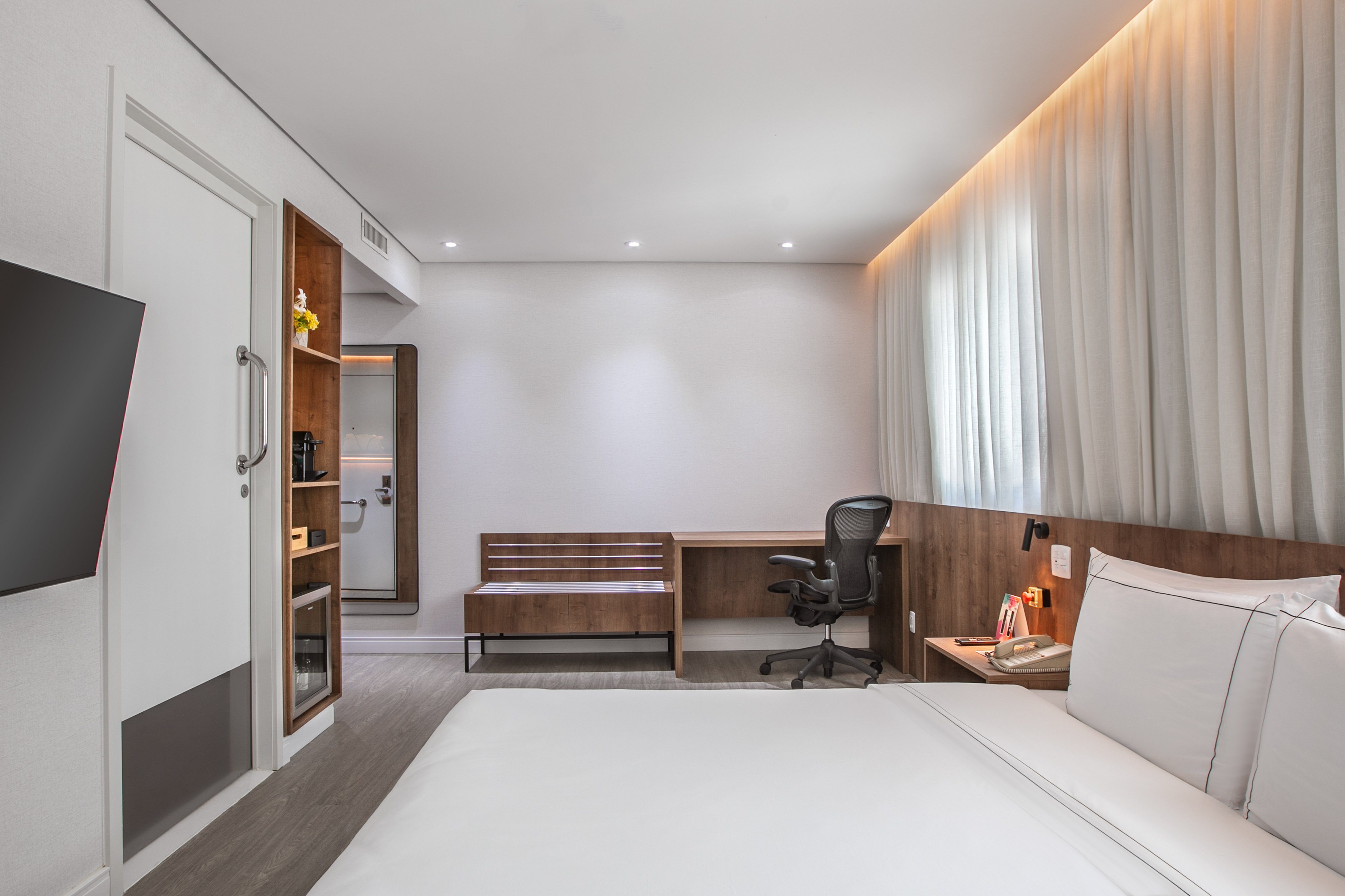 Modern hotel room with white bed, wooden desk, workspace, and backlit curtains.
